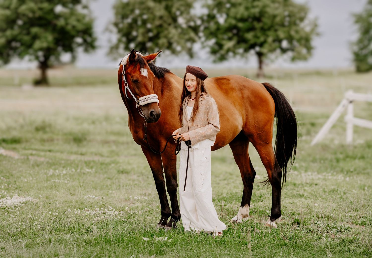 Young woman in white dress and beret stands beside a chestnut bay horse in a green pasture.