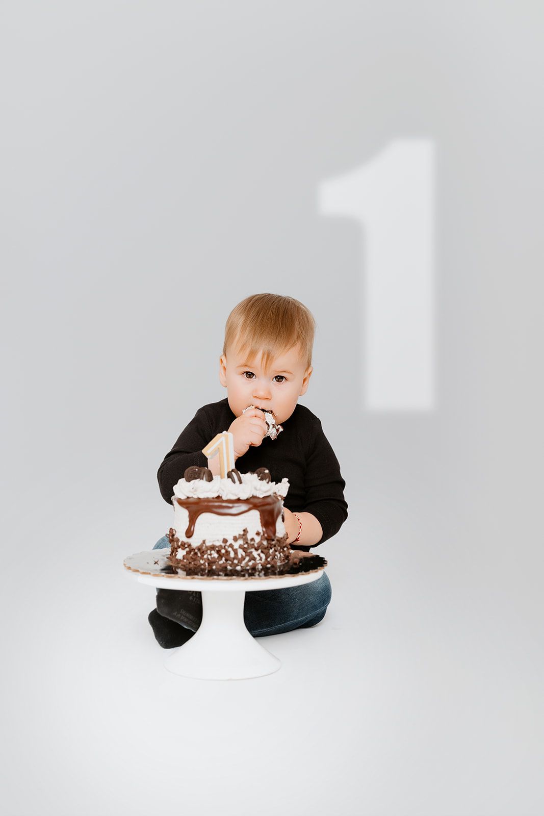 A toddler in a black outfit sits on the floor, eating from a chocolate birthday cake with a number 1 on the wall.