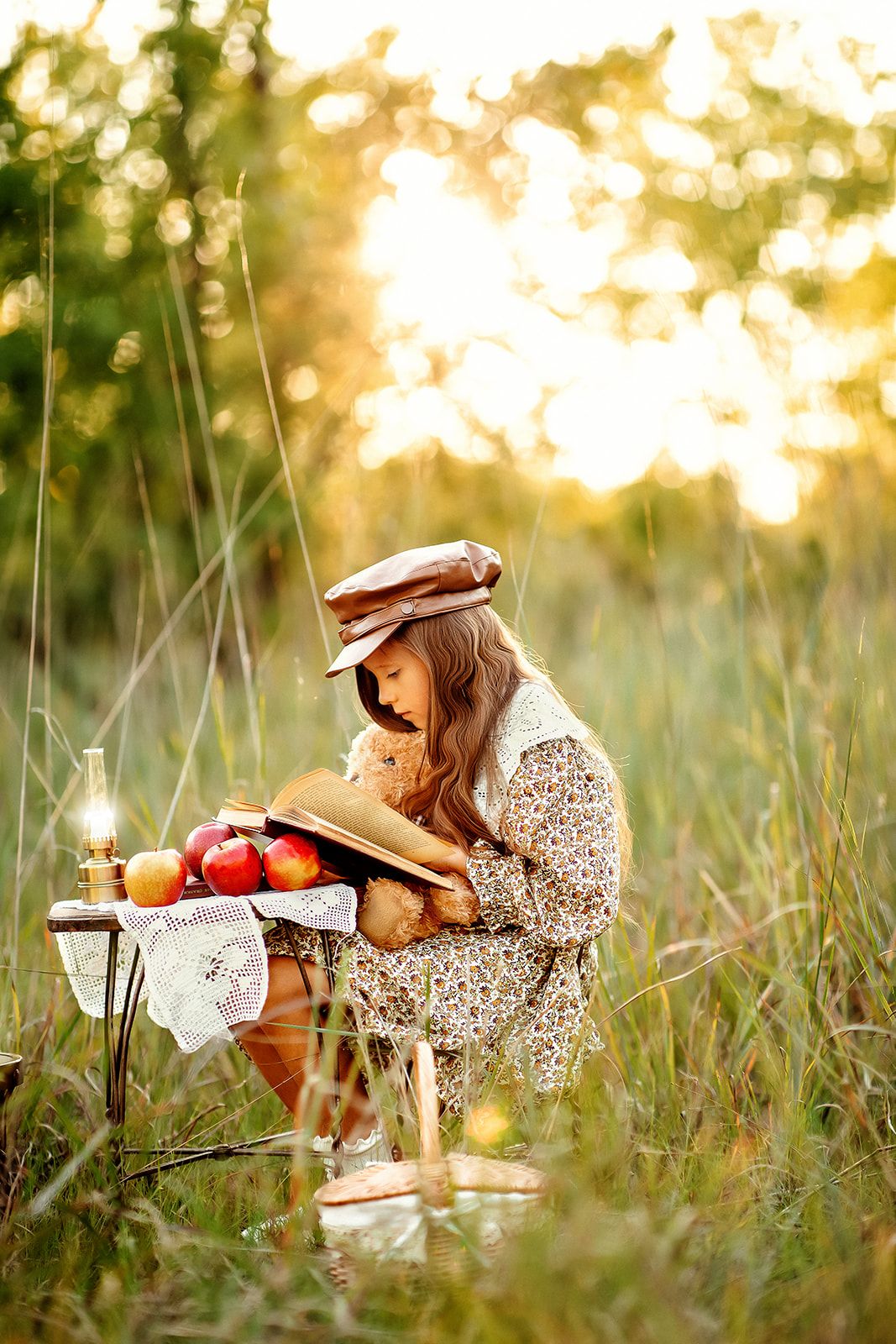 A young girl in a floral dress and cap reads a book outdoors at sunset near apples and a lantern.