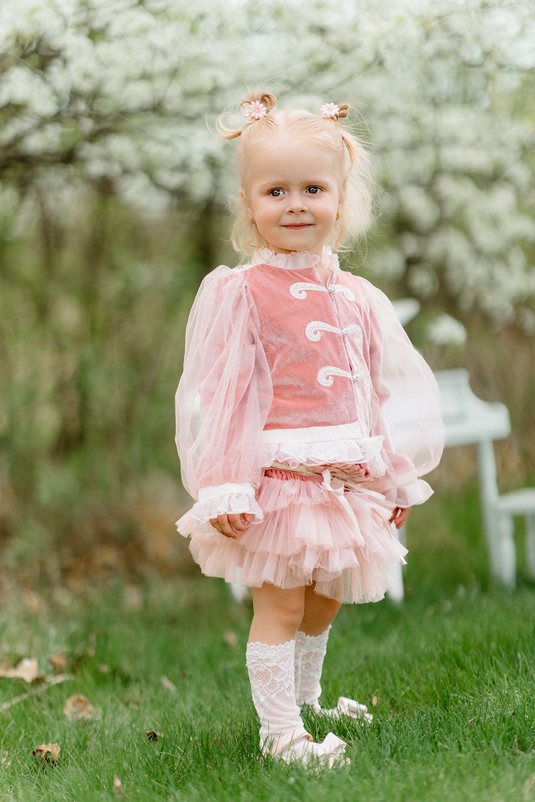 A cute toddler girl in a pink tutu dress and lace socks stands on green grass near blooming white trees.