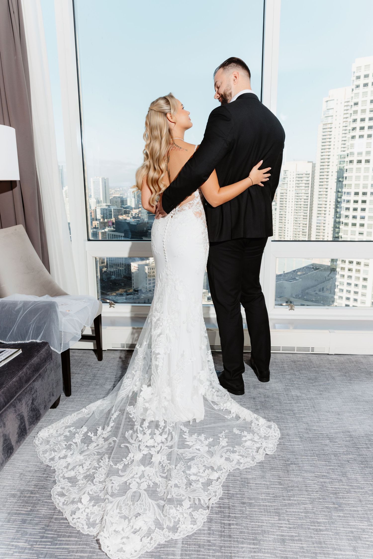Bride in lace mermaid gown with long train and groom in black suit embrace facing city skyline through large windows.