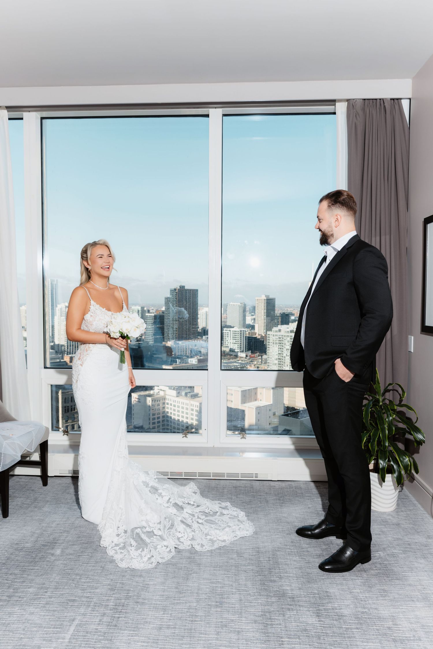 Bride laughing joyfully holding white bouquet during first look with groom in city hotel room.