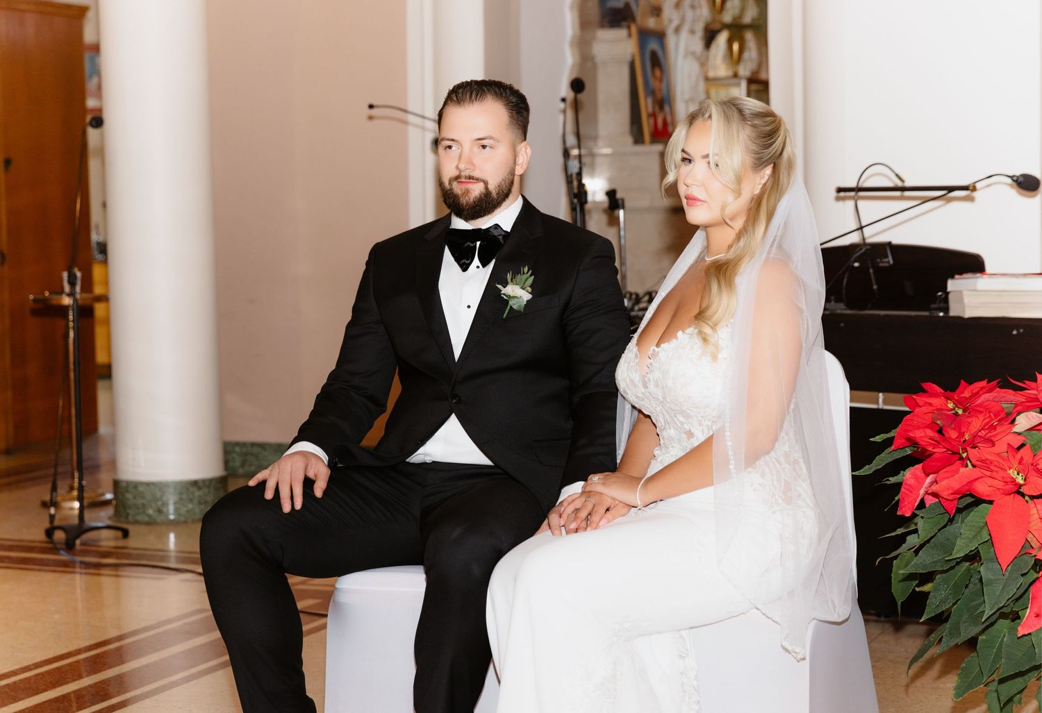 Bride and groom sit together during church wedding ceremony surrounded by red poinsettias and holiday decorations.