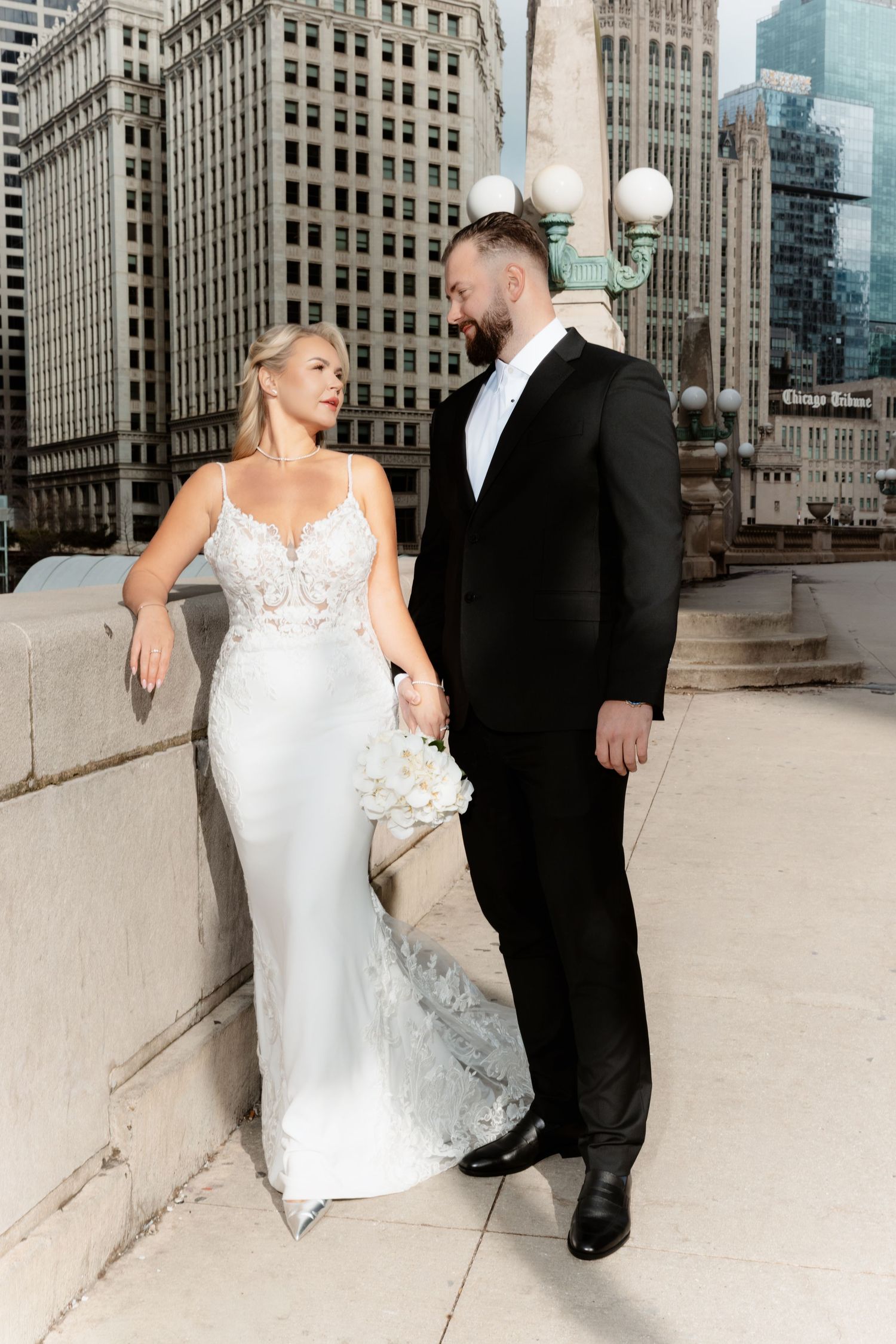 Bride in lace gown and groom in tuxedo share romantic moment on Chicago Riverwalk bridge.
