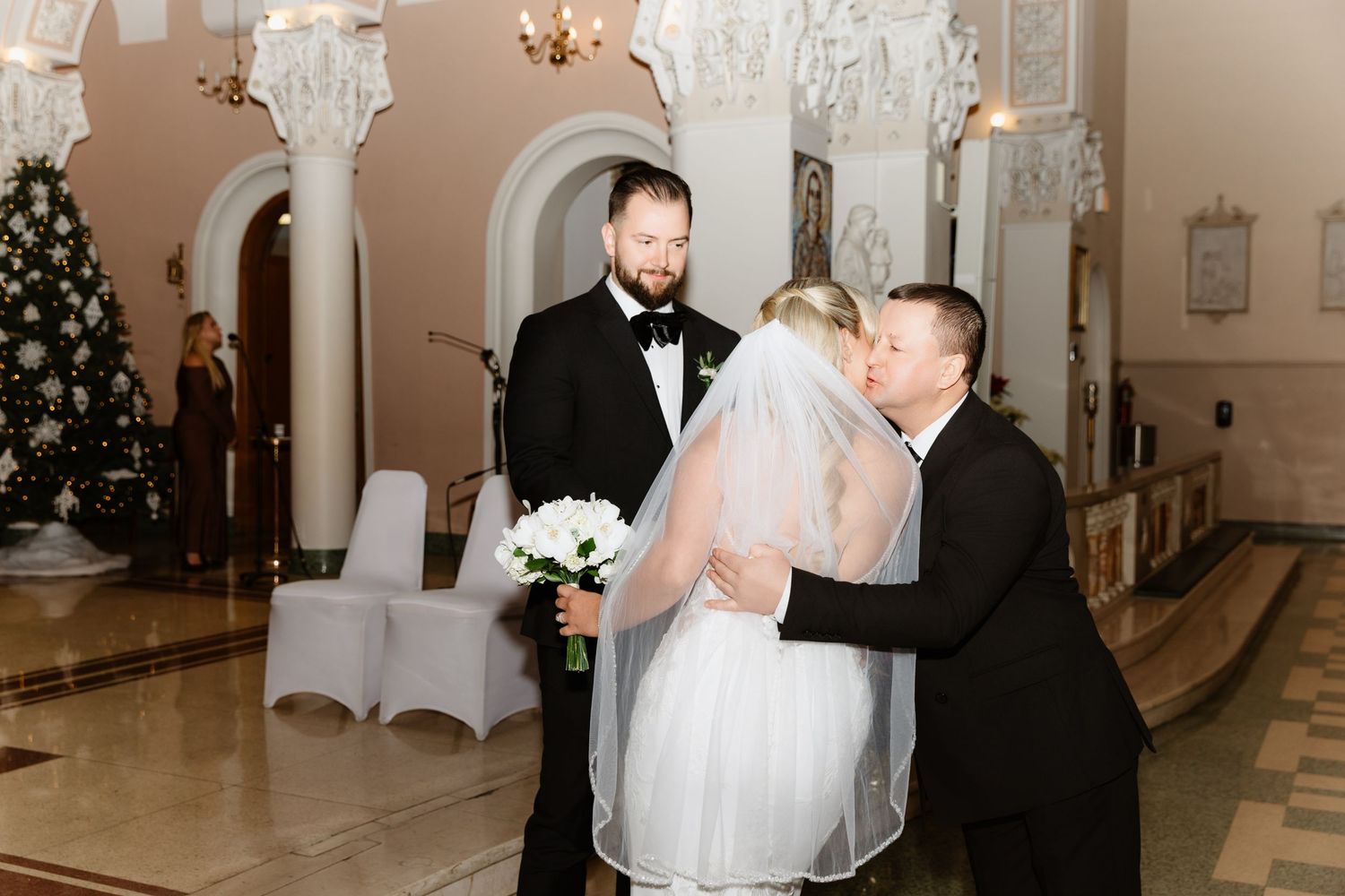 Groom embraces bride in white lace dress as officiant stands nearby in Christmas decorated church.