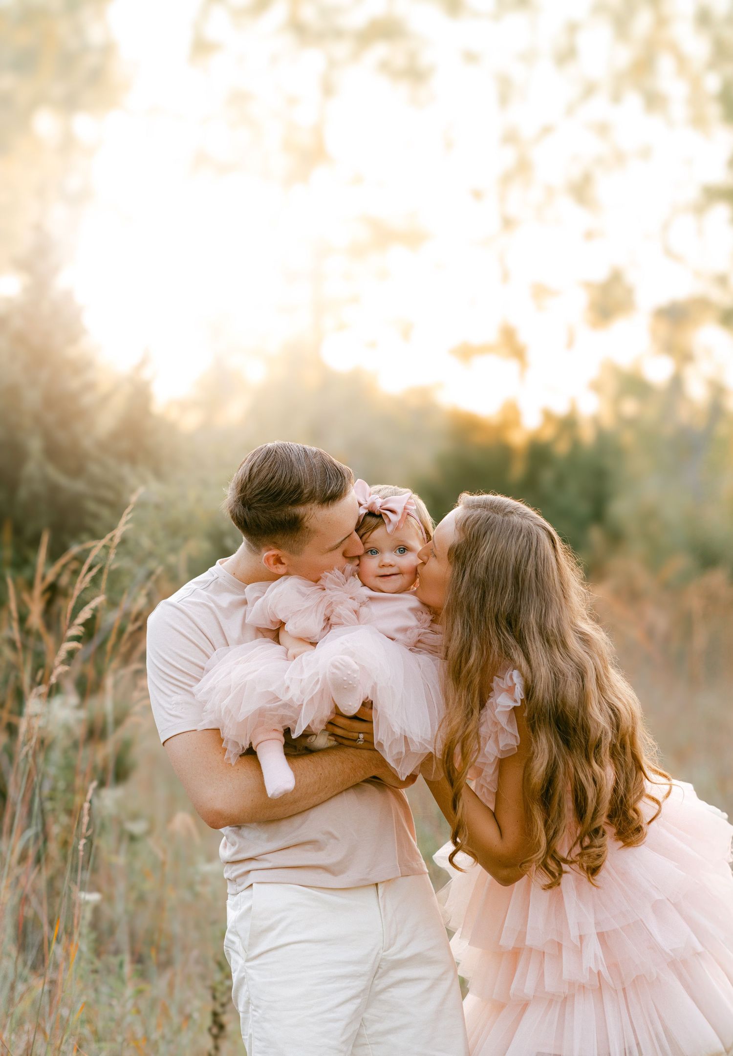 Parents kissing their smiling baby girl in pink tulle dress during golden hour outdoor session.
