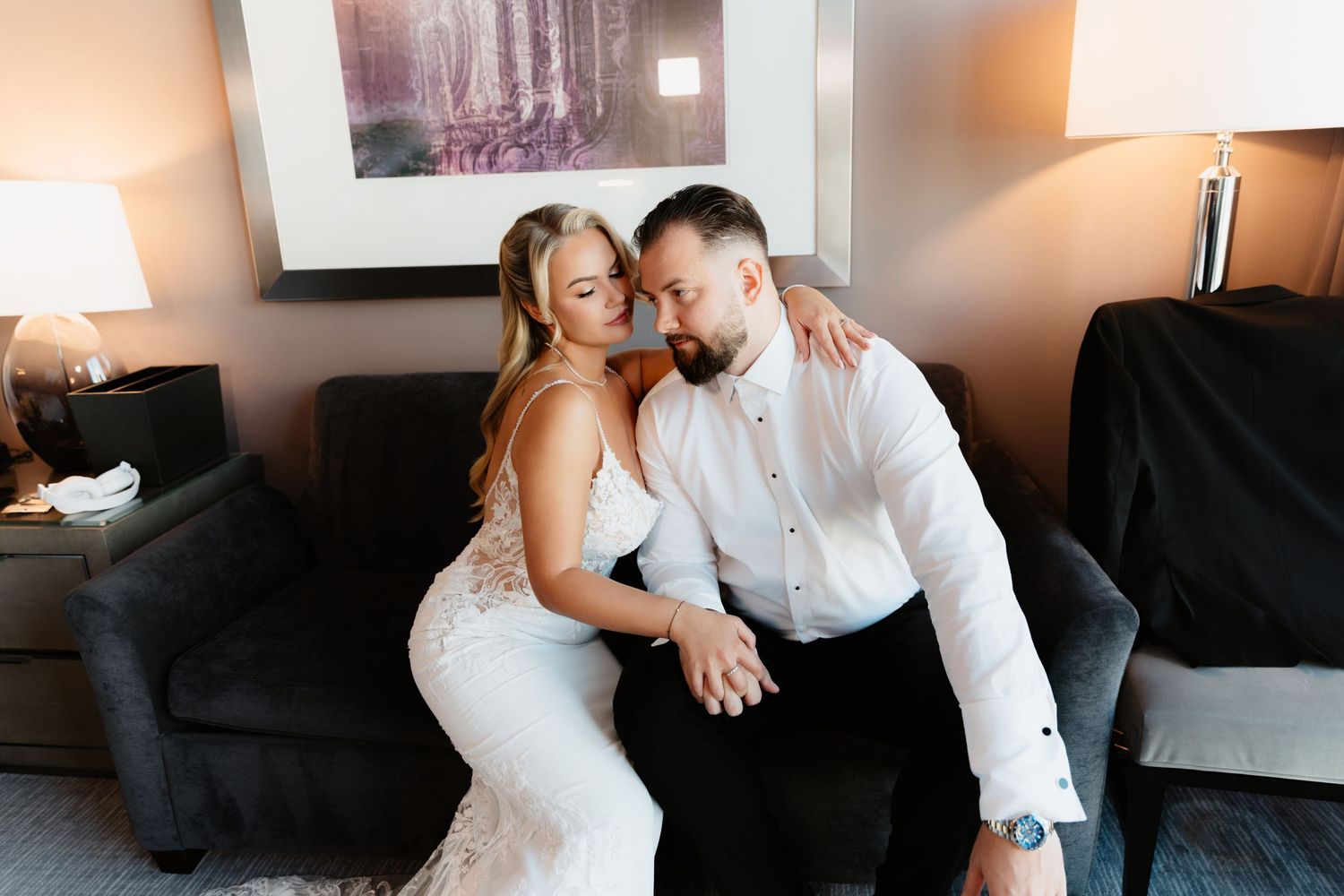Bride in lace wedding dress and groom in white shirt share a romantic moment on sofa.