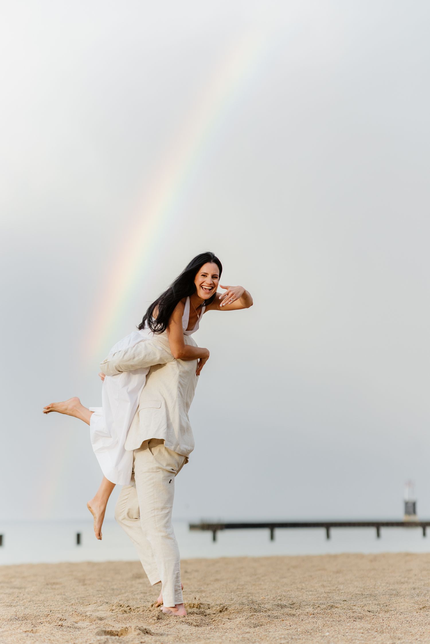 A joyful couple on a sandy beach with a rainbow, man giving woman a piggyback ride, both dressed in white.