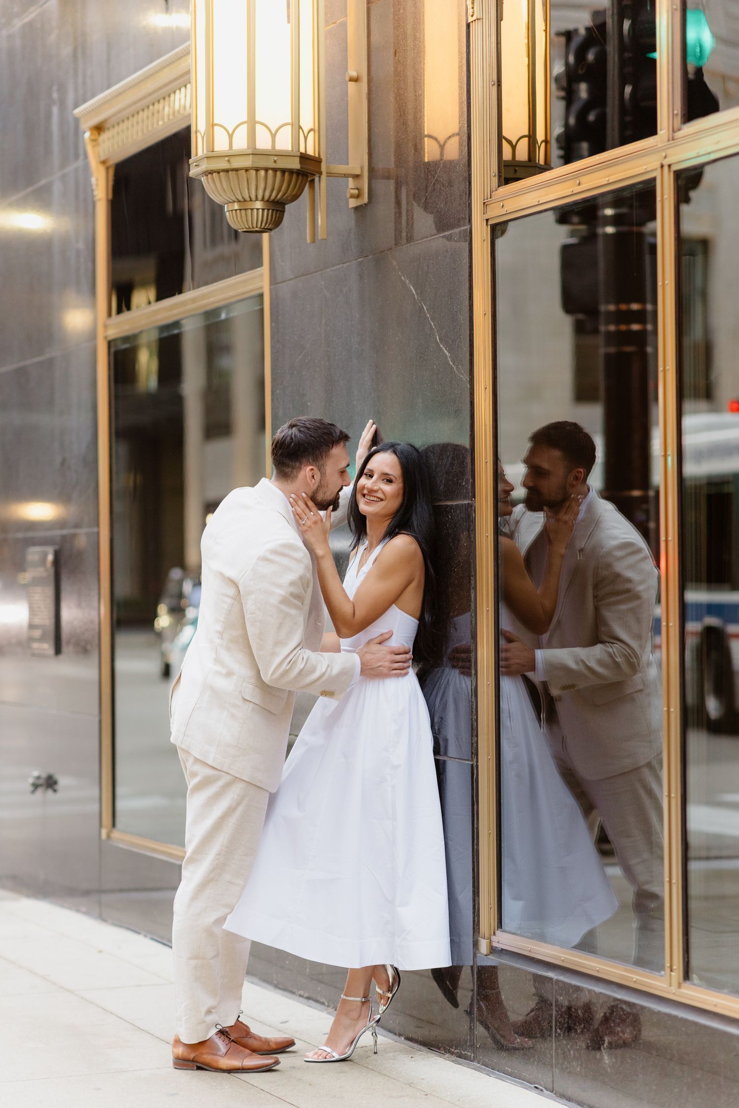 Engaged couple sharing a romantic moment against a gold-framed building facade on a city sidewalk.