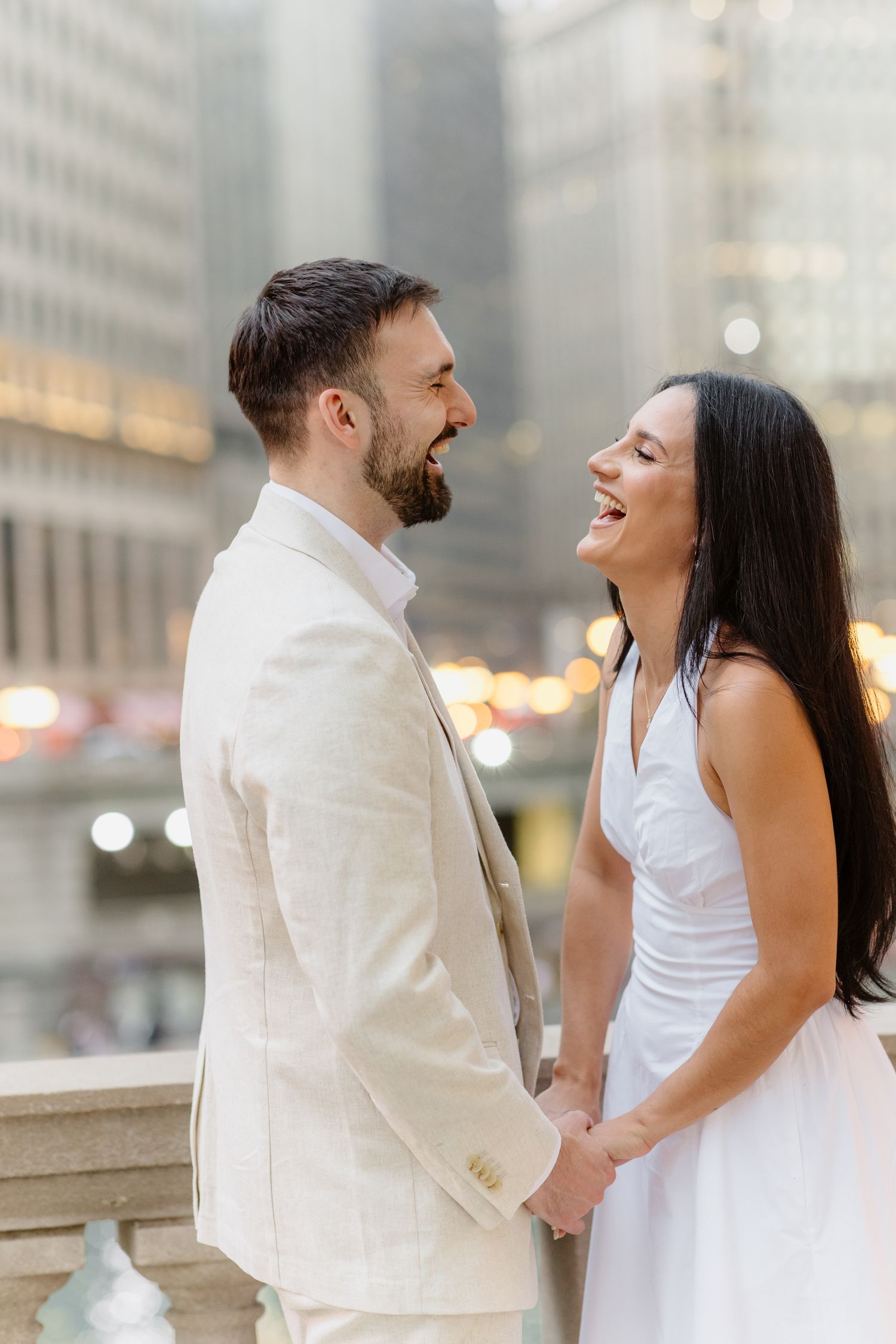 Laughing couple in white attire holding hands facing each other with city buildings behind.
