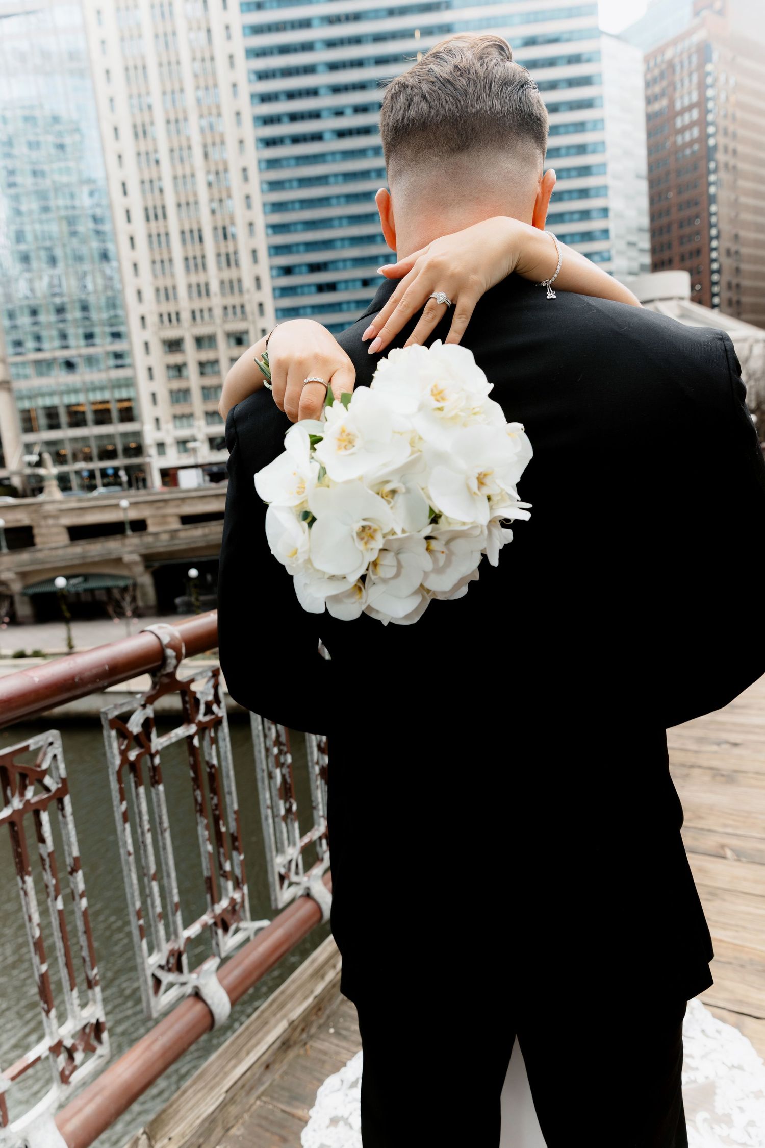 Bride embracing groom from behind on bridge holding white orchid bouquet with city skyline backdrop.