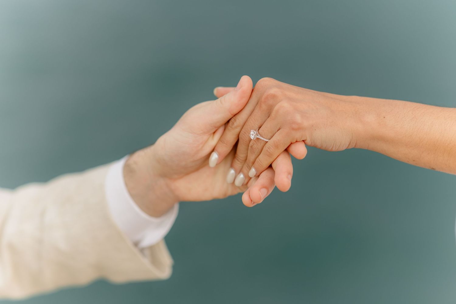 Close-up of couple holding hands showing engagement ring against teal background.