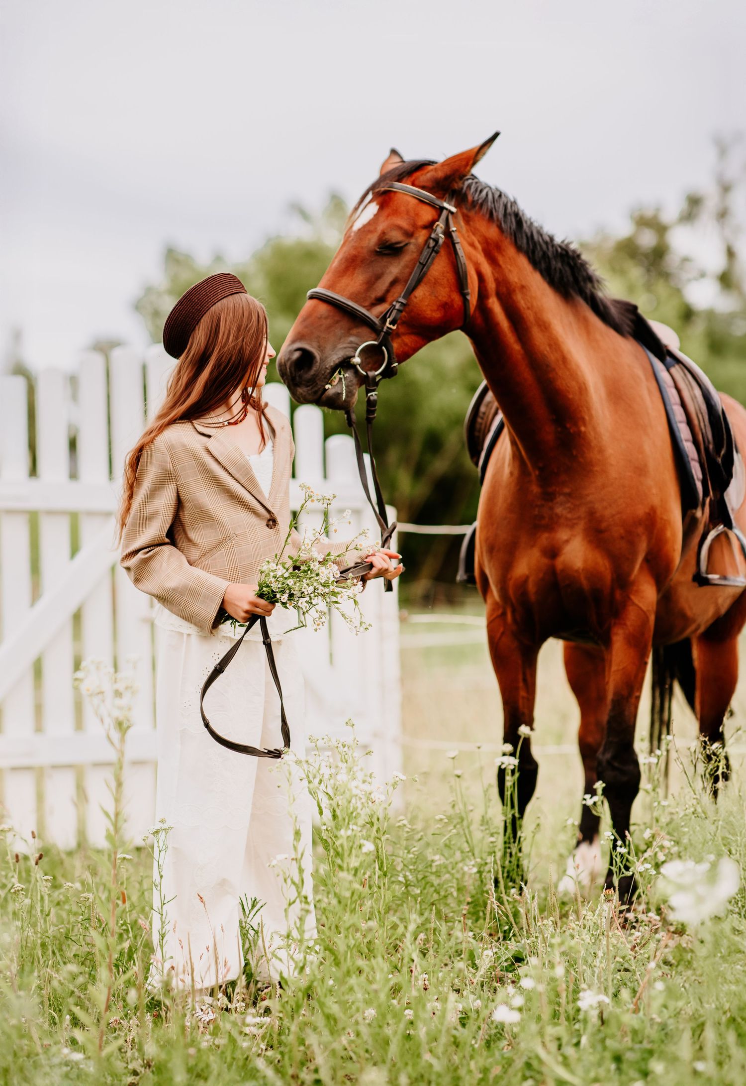 Young woman in beige coat and hat faces a chestnut horse holding wildflowers by a white picket fence.