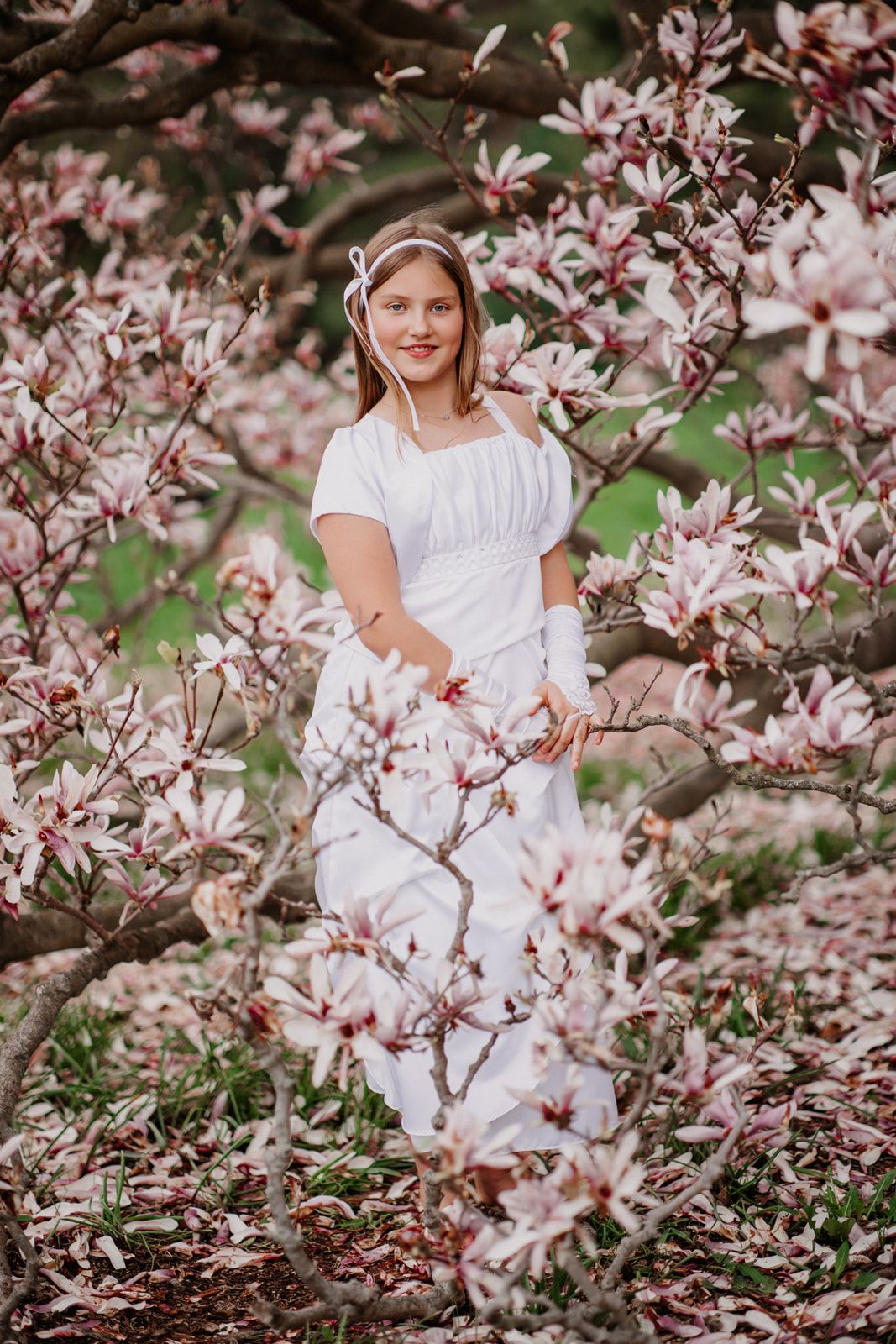 Young woman in white dress standing among blooming pink magnolia tree branches in a lush garden.