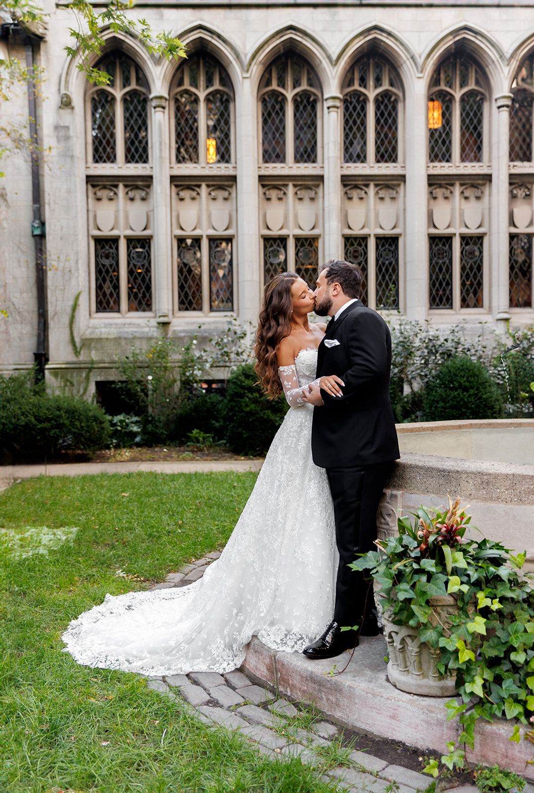 Bride and groom share a romantic kiss in a lace wedding gown and tuxedo outside a Gothic stone church.