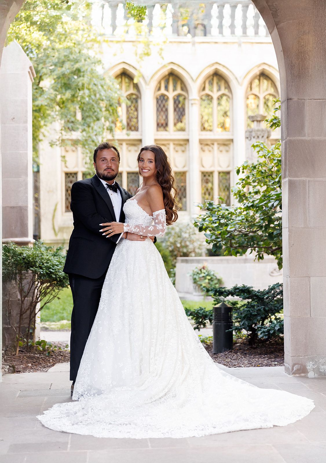 Elegant bride and groom posing in white lace wedding gown and black tuxedo under gothic stone archway.