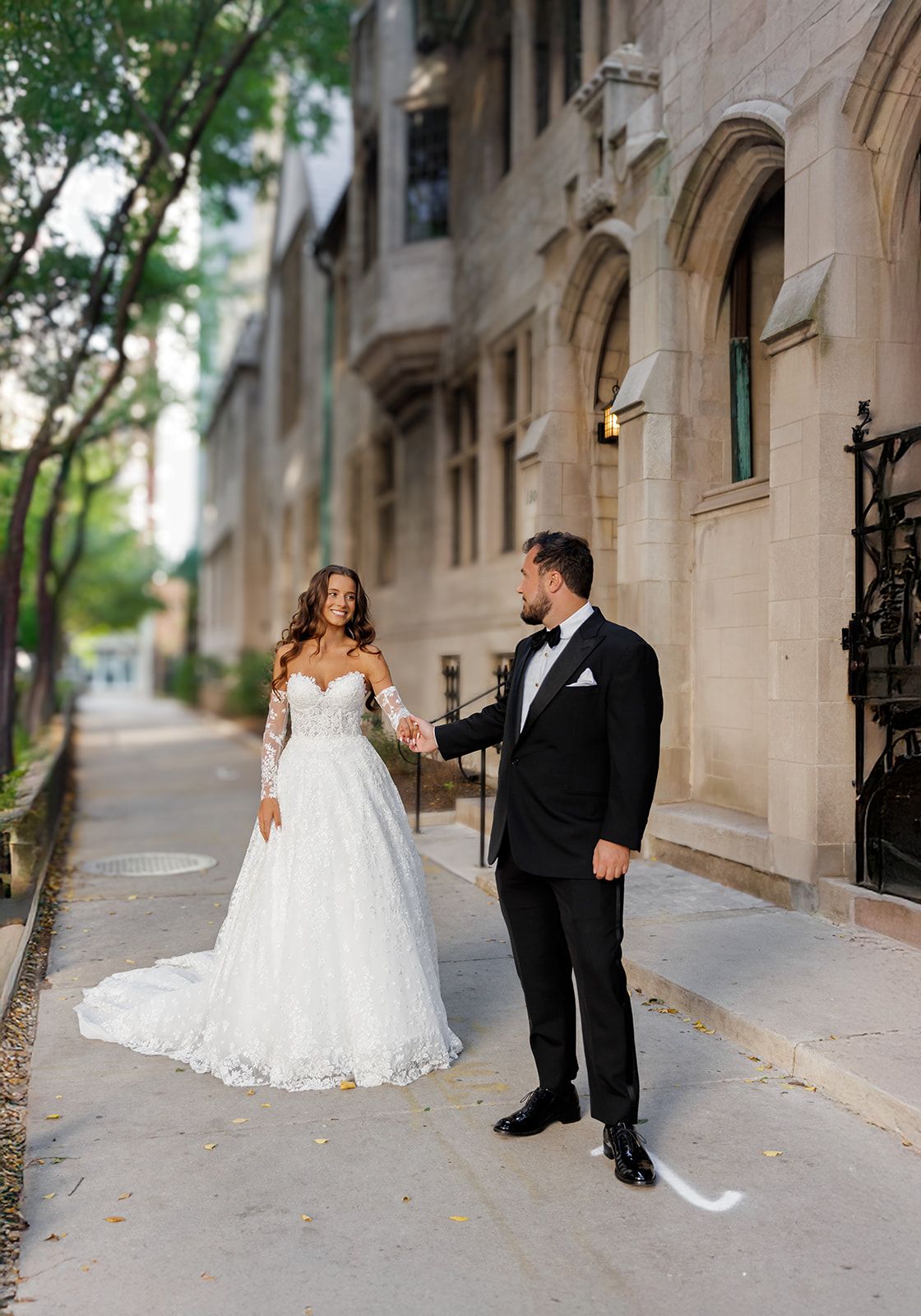 Bride in lace off-shoulder gown and groom in tuxedo hold hands on a tree-lined sidewalk outside a stone building.