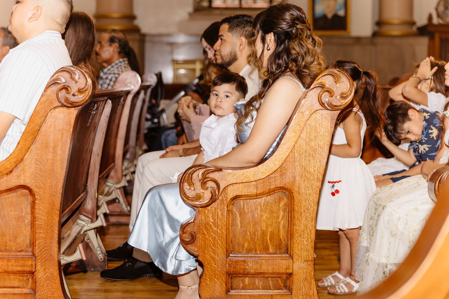Family sitting in wooden church pews during a religious ceremony, with a mother holding a baby dressed in white.