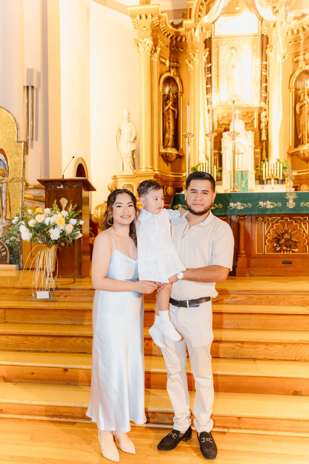 Parents in elegant attire hold baby in white baptism outfit inside ornate Catholic church.