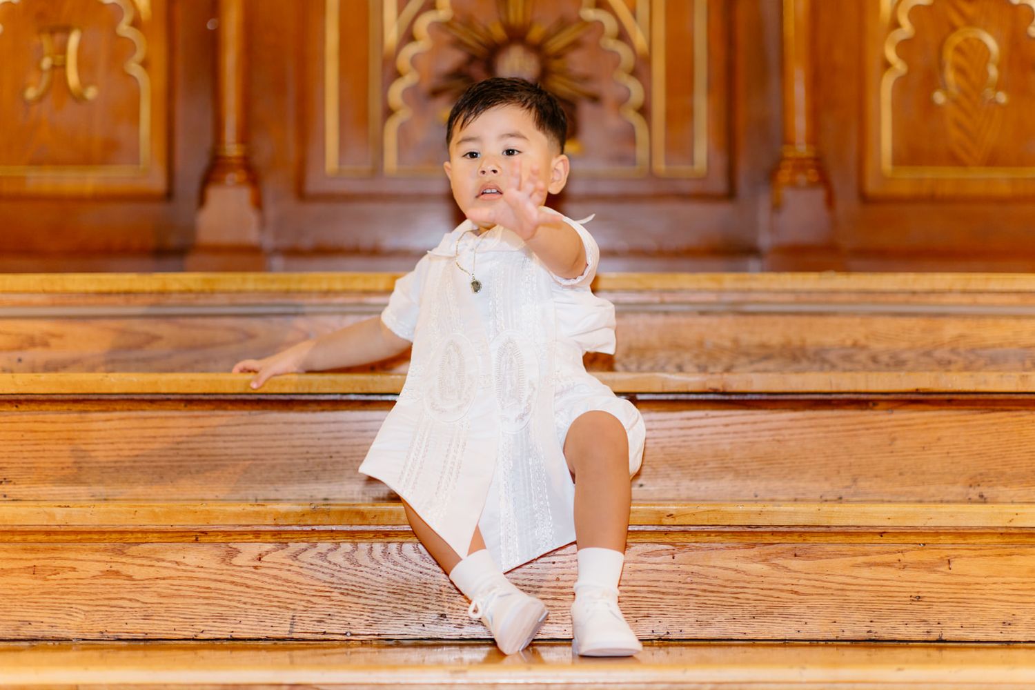 Cute toddler in white baptism outfit sitting on wooden church steps reaching forward.