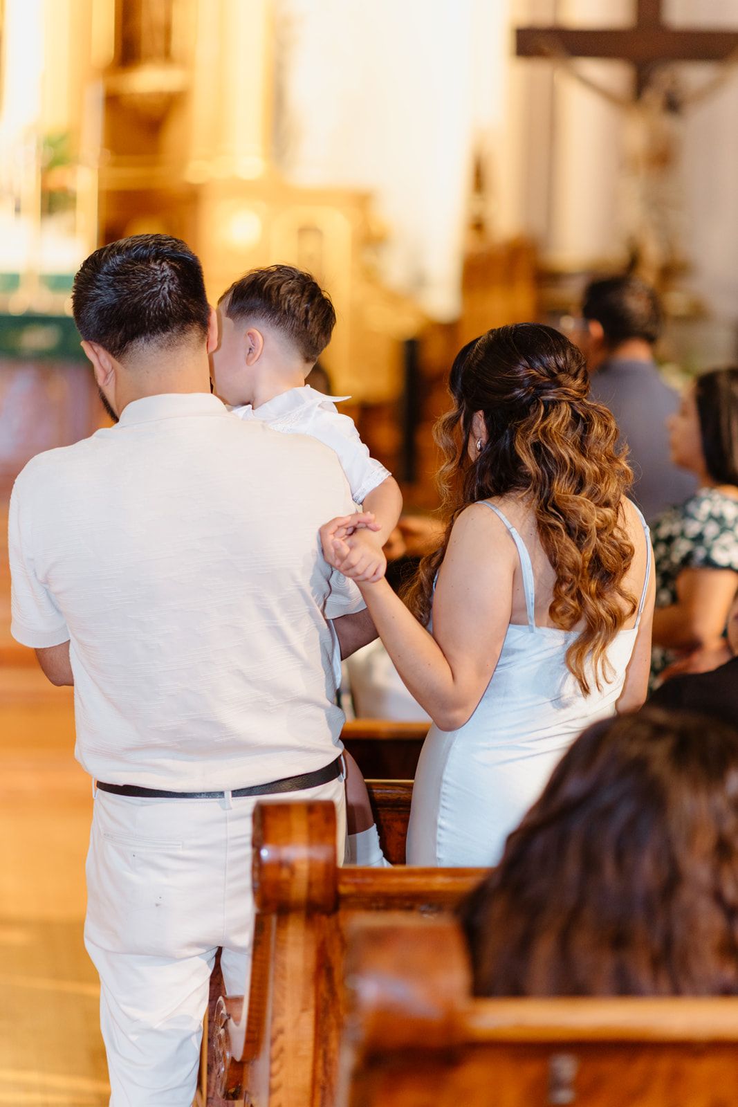 Family in white attire standing together during a church ceremony, viewed from behind near wooden pews.