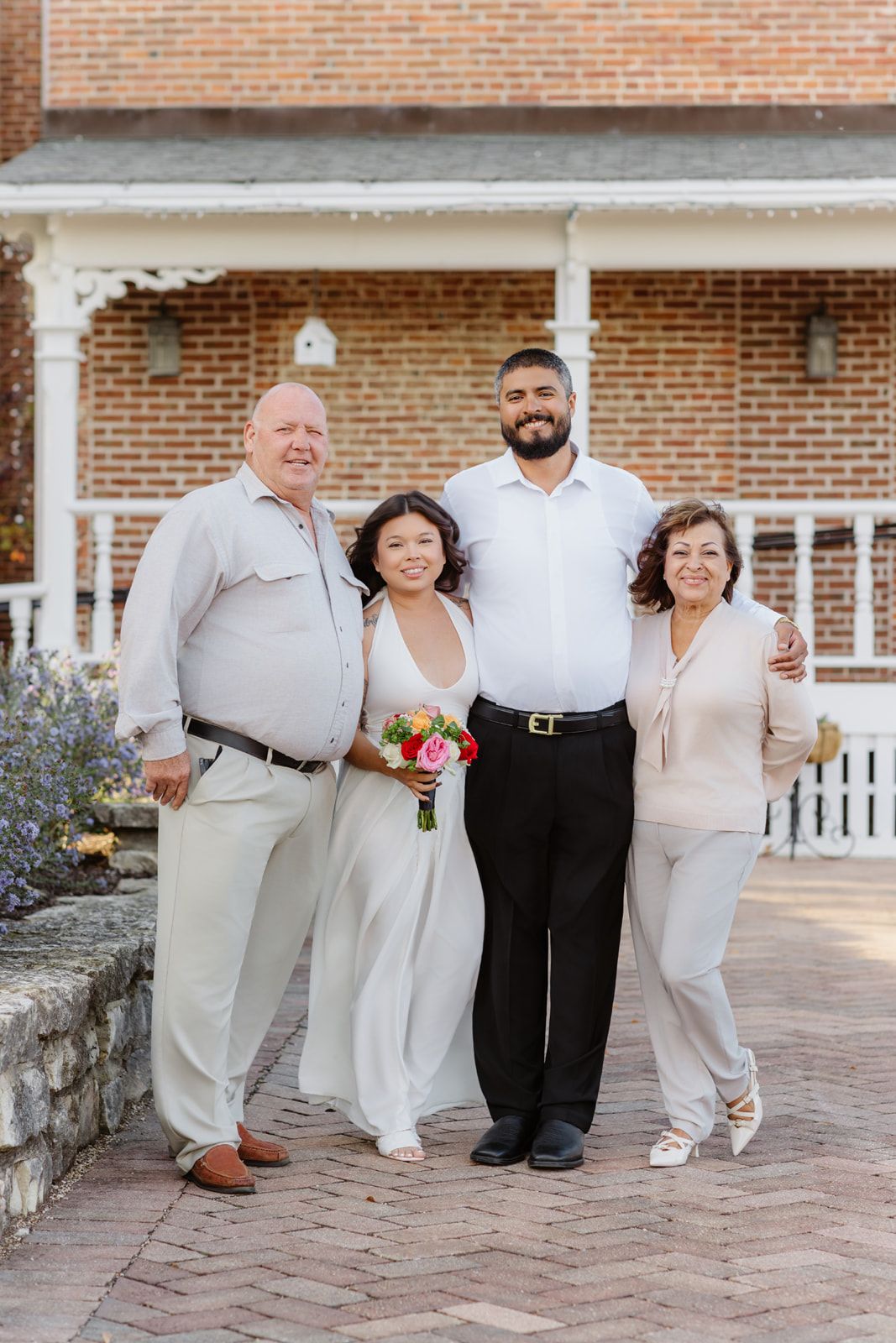 Bride and groom smile with older couple in front of brick building with white porch.
