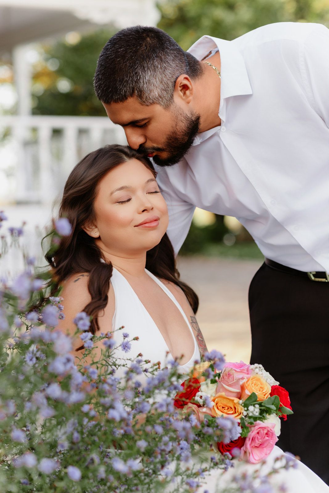 Groom tenderly kisses bride on forehead as she holds colorful bouquet among purple flowers.