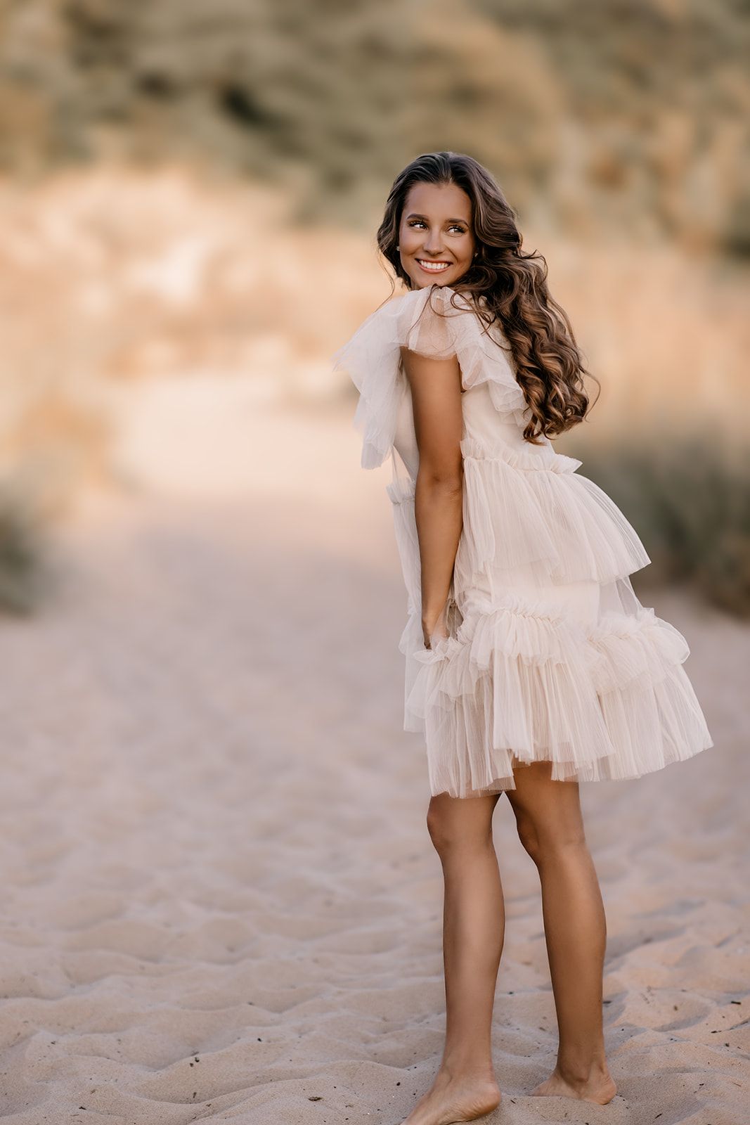 Smiling woman in elegant white tulle dress standing barefoot on sandy beach path at golden hour.