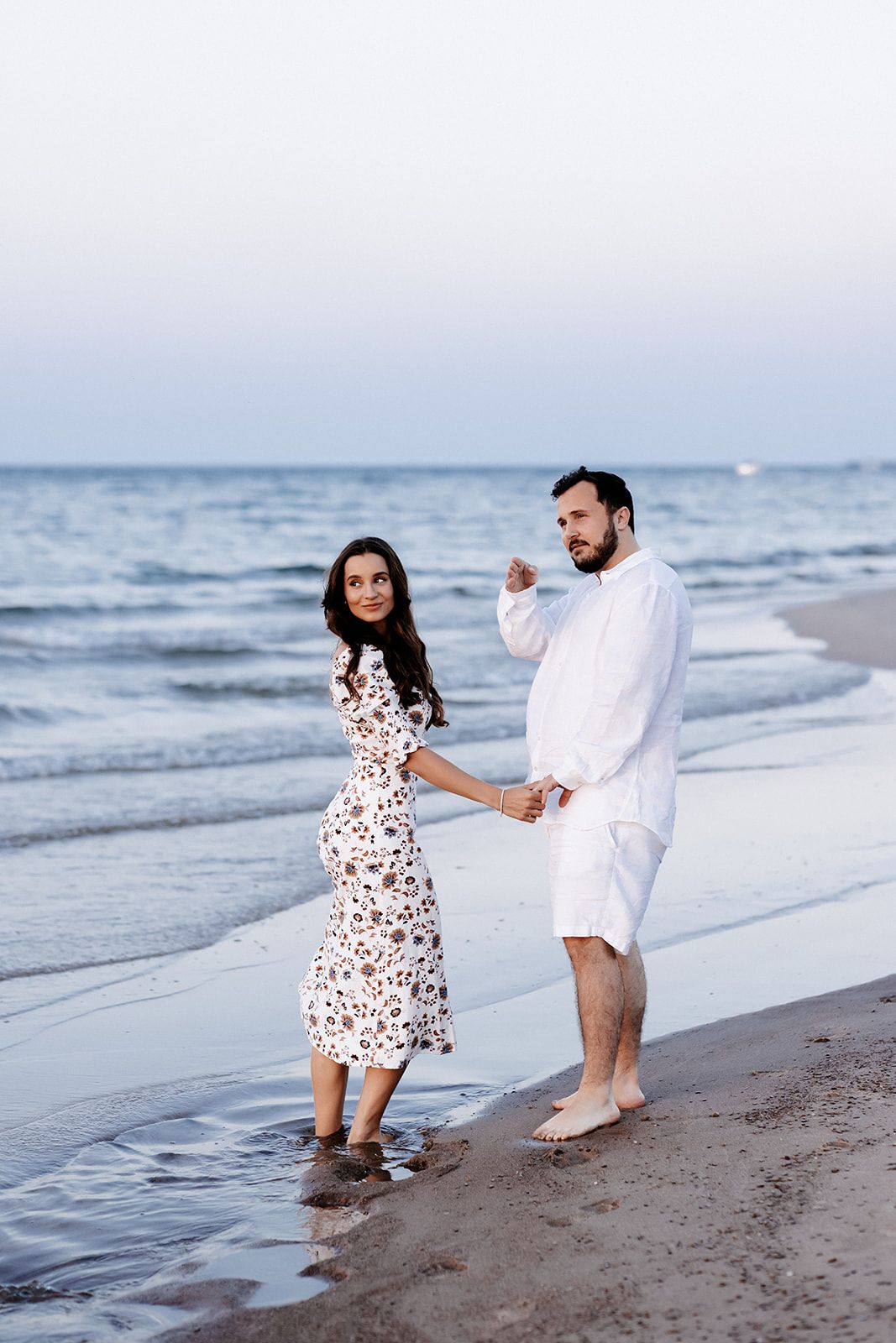 Couple in white outfits holding hands on a sandy beach at dusk with gentle ocean waves in the background.