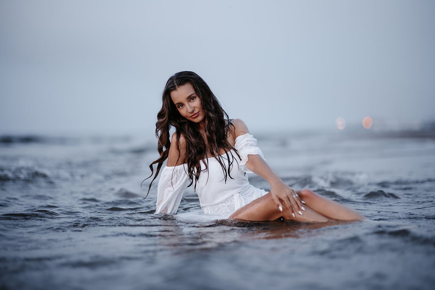 Young woman in white off-shoulder dress sitting in shallow ocean waves with wet hair on an overcast day.