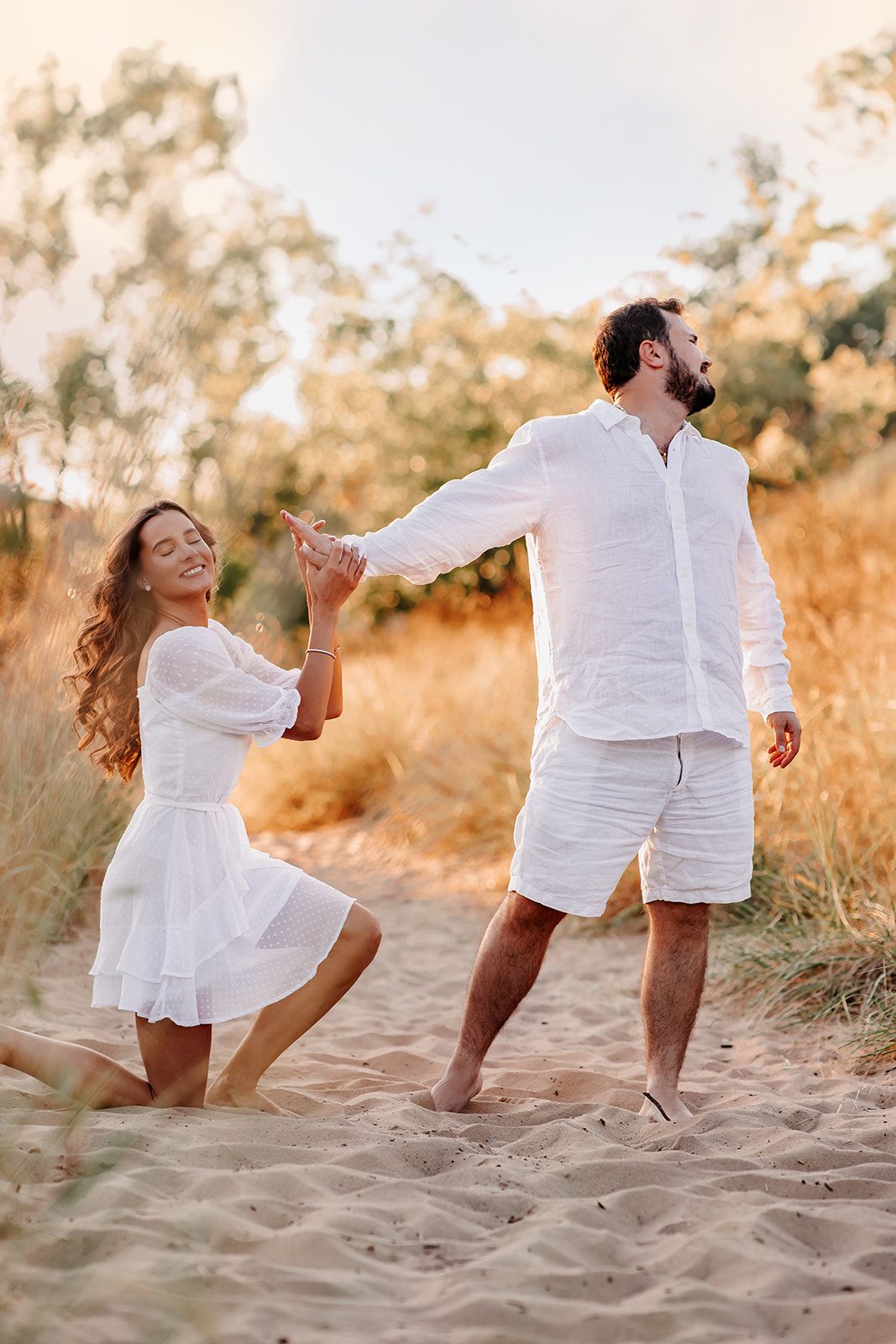 Couple in white outfits dancing playfully on sandy beach dunes at golden sunset.