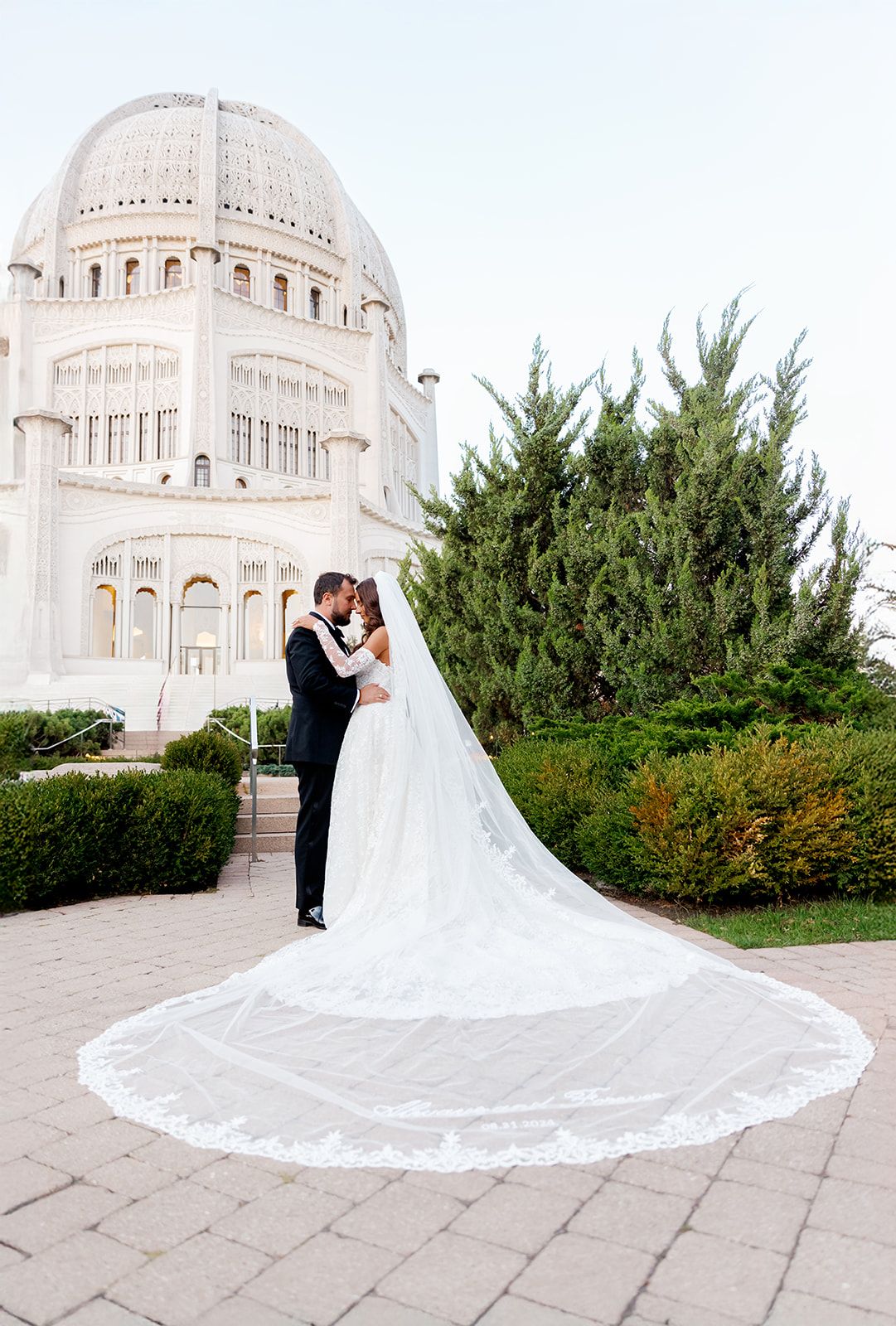 Bride with dramatic cathedral lace veil kissing groom outside white Bahai Temple.