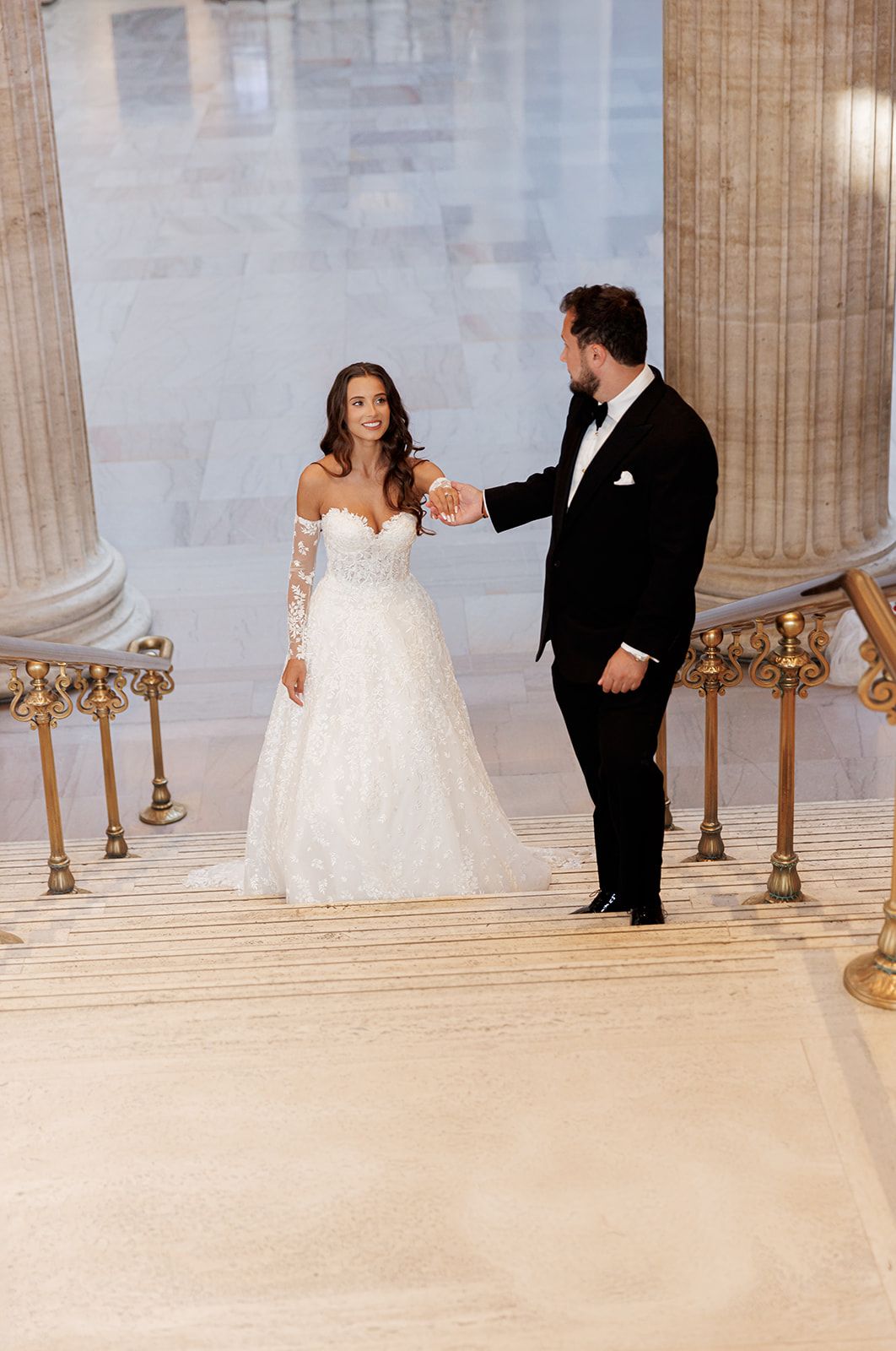 Groom holds bride's hand on ornate marble steps, bride wearing off-shoulder lace ball gown.