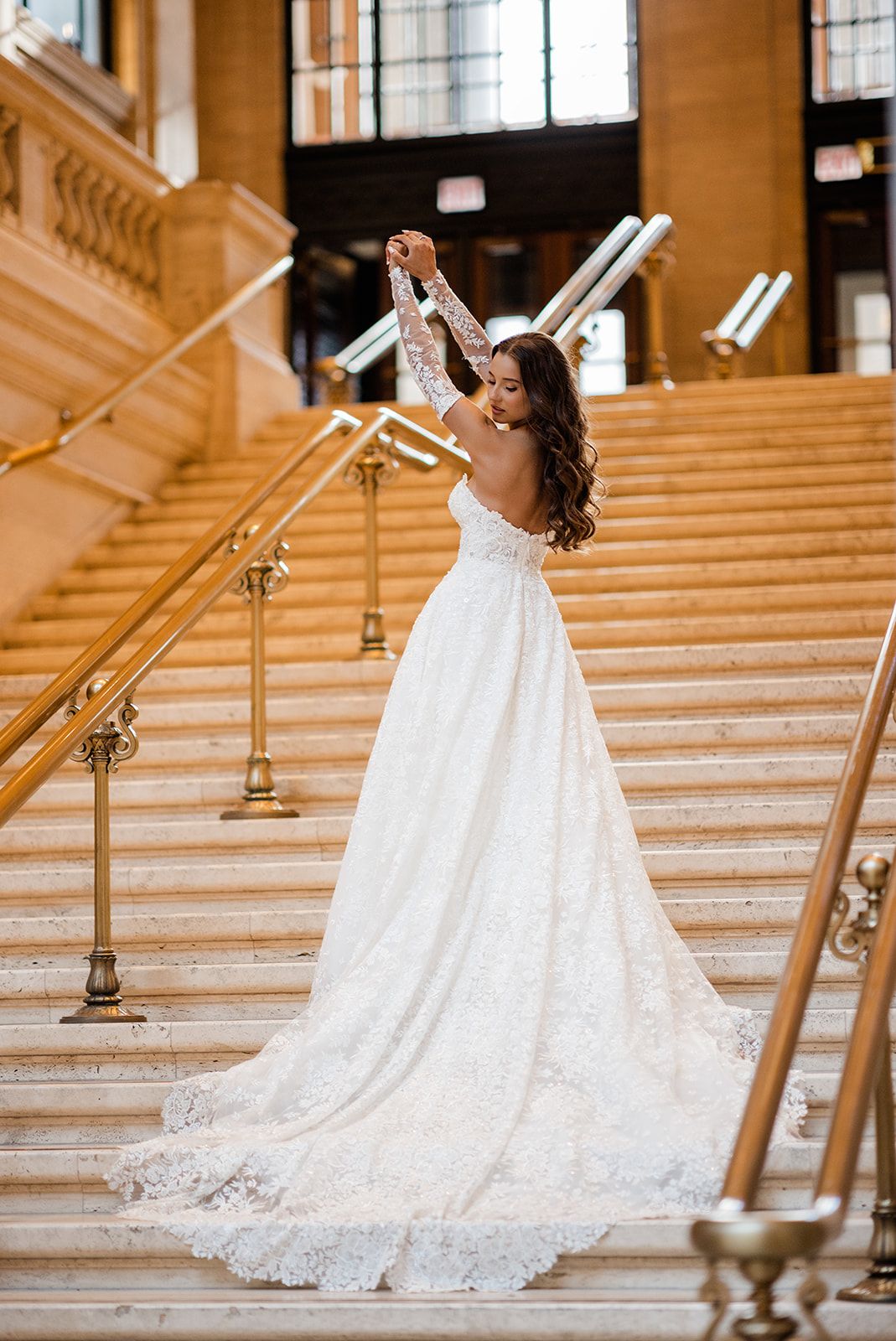 Bride in lace wedding gown with long sleeves poses on grand staircase with gold railings in elegant venue.
