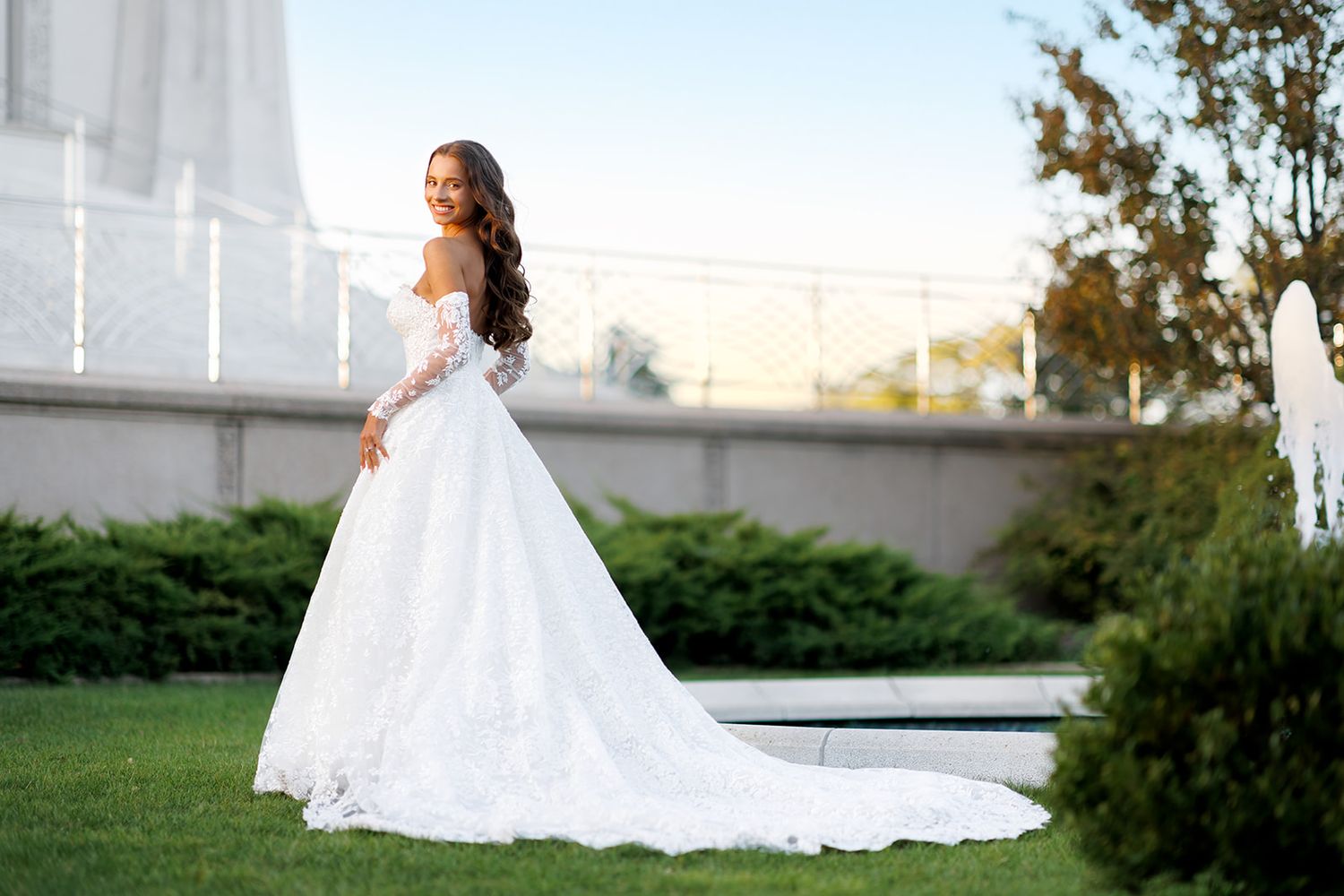Bride in elegant white lace ball gown with long train poses gracefully in garden setting.