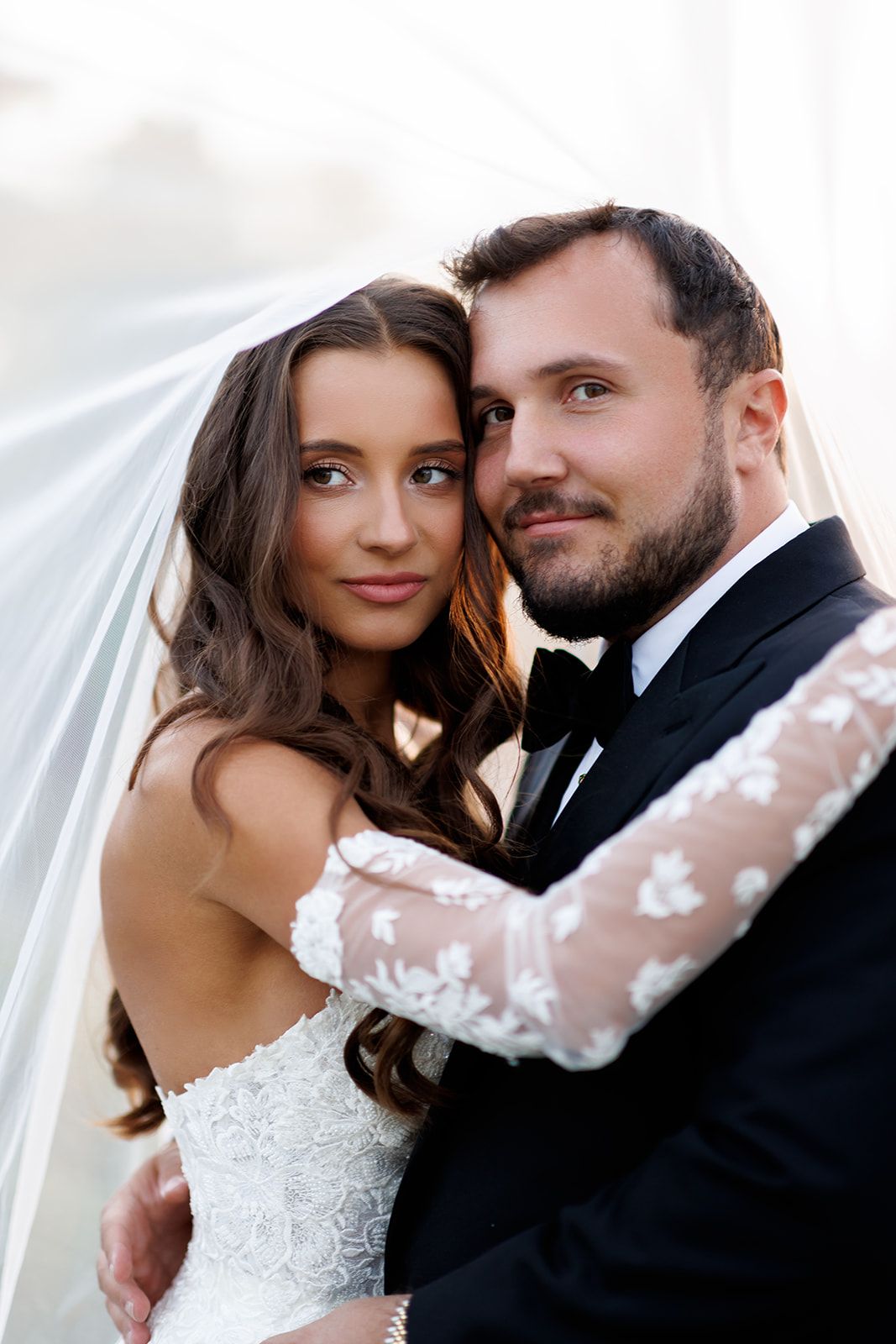 Elegant bride and groom embrace closely under a flowing veil in a romantic wedding portrait.
