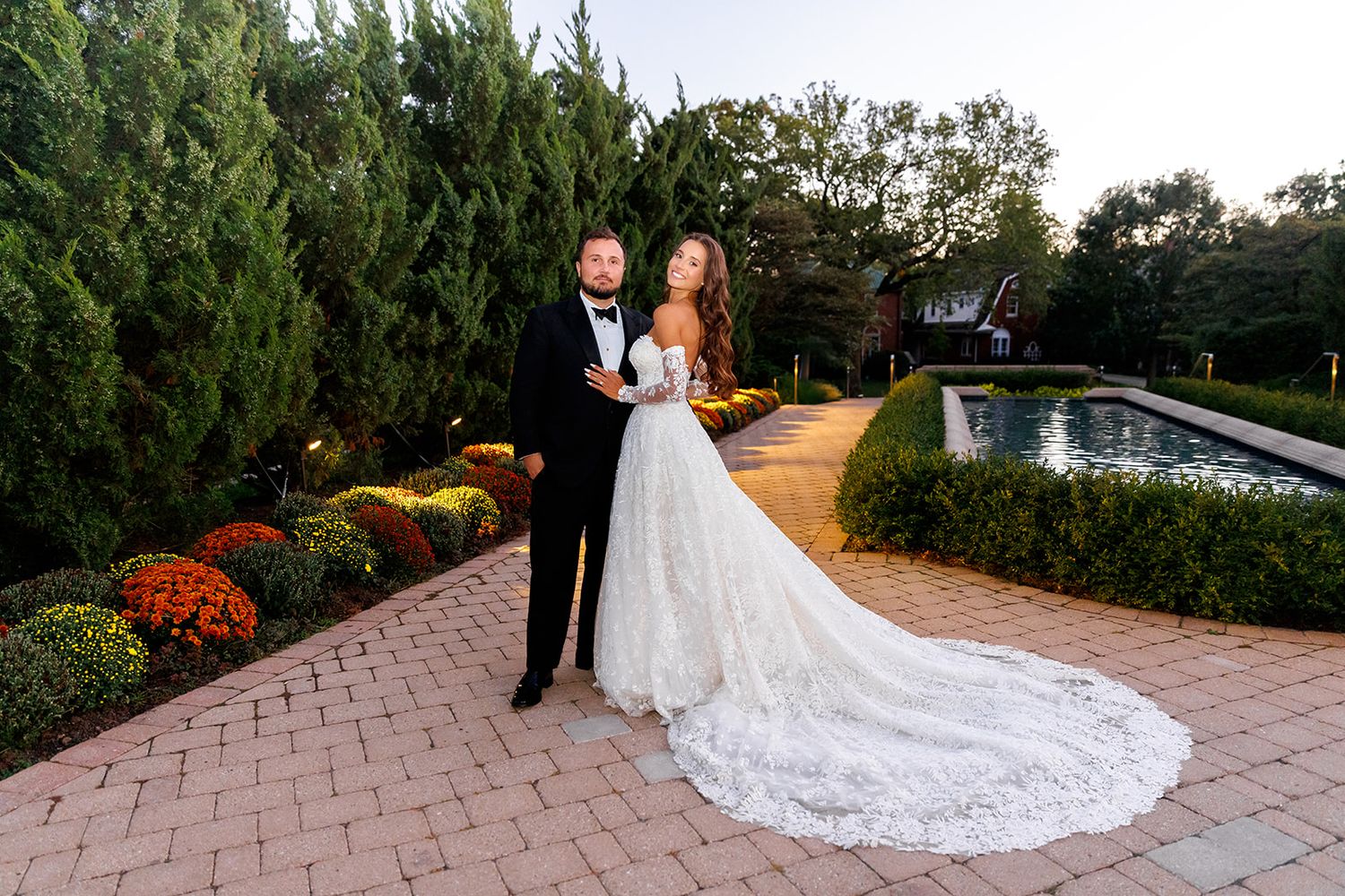 Bride and groom pose elegantly on brick path surrounded by lush gardens and a pond at dusk.