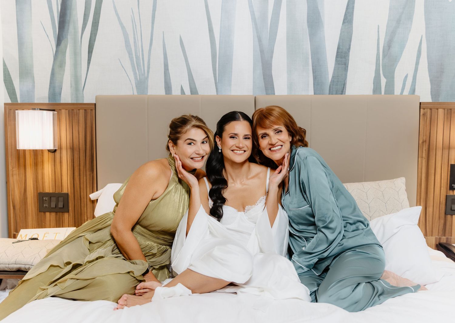 Bride in white robe poses joyfully with two women in green and teal dresses on white hotel bed before wedding.