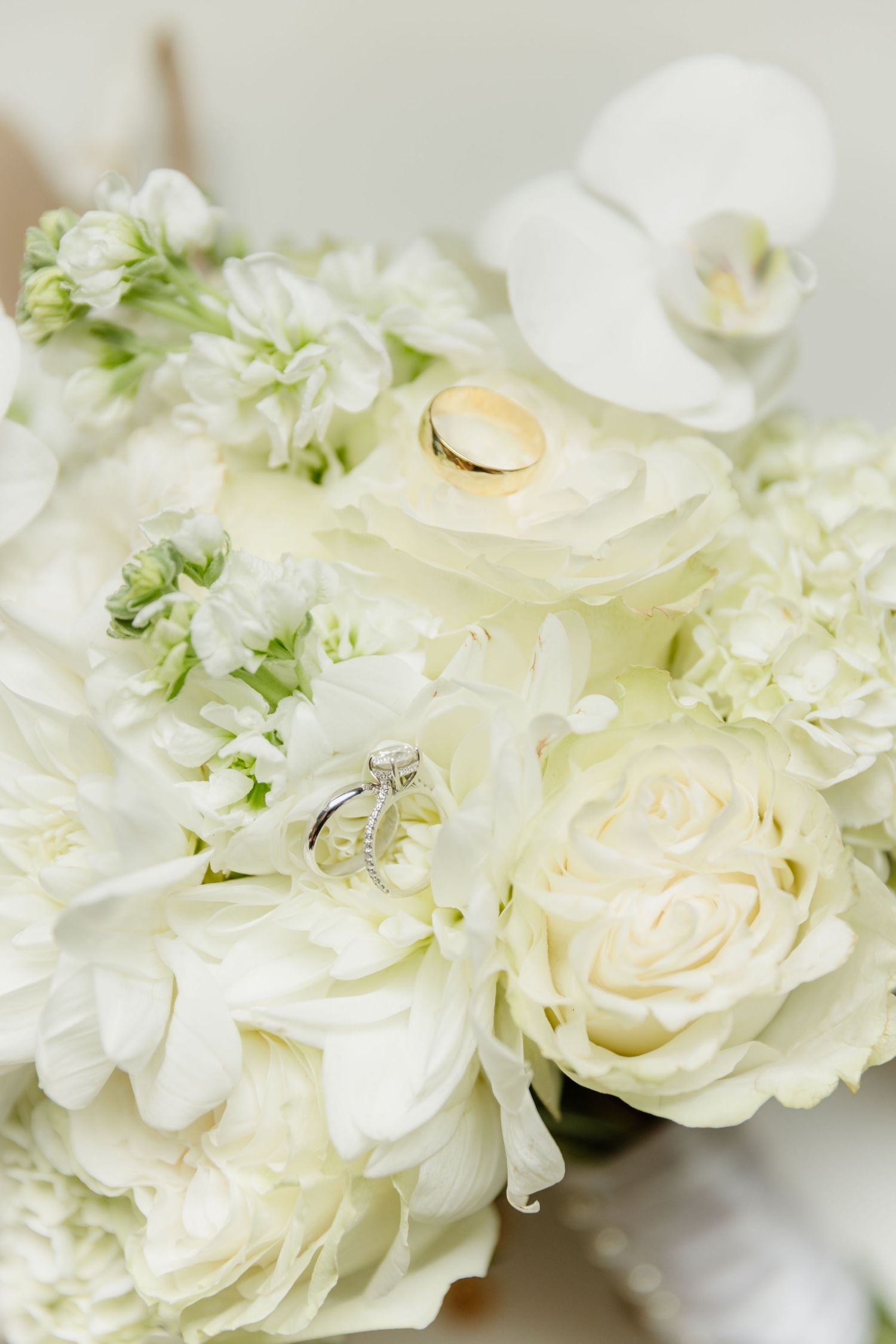 White wedding bouquet with roses, peonies, and orchids displaying a diamond engagement ring and gold wedding band.
