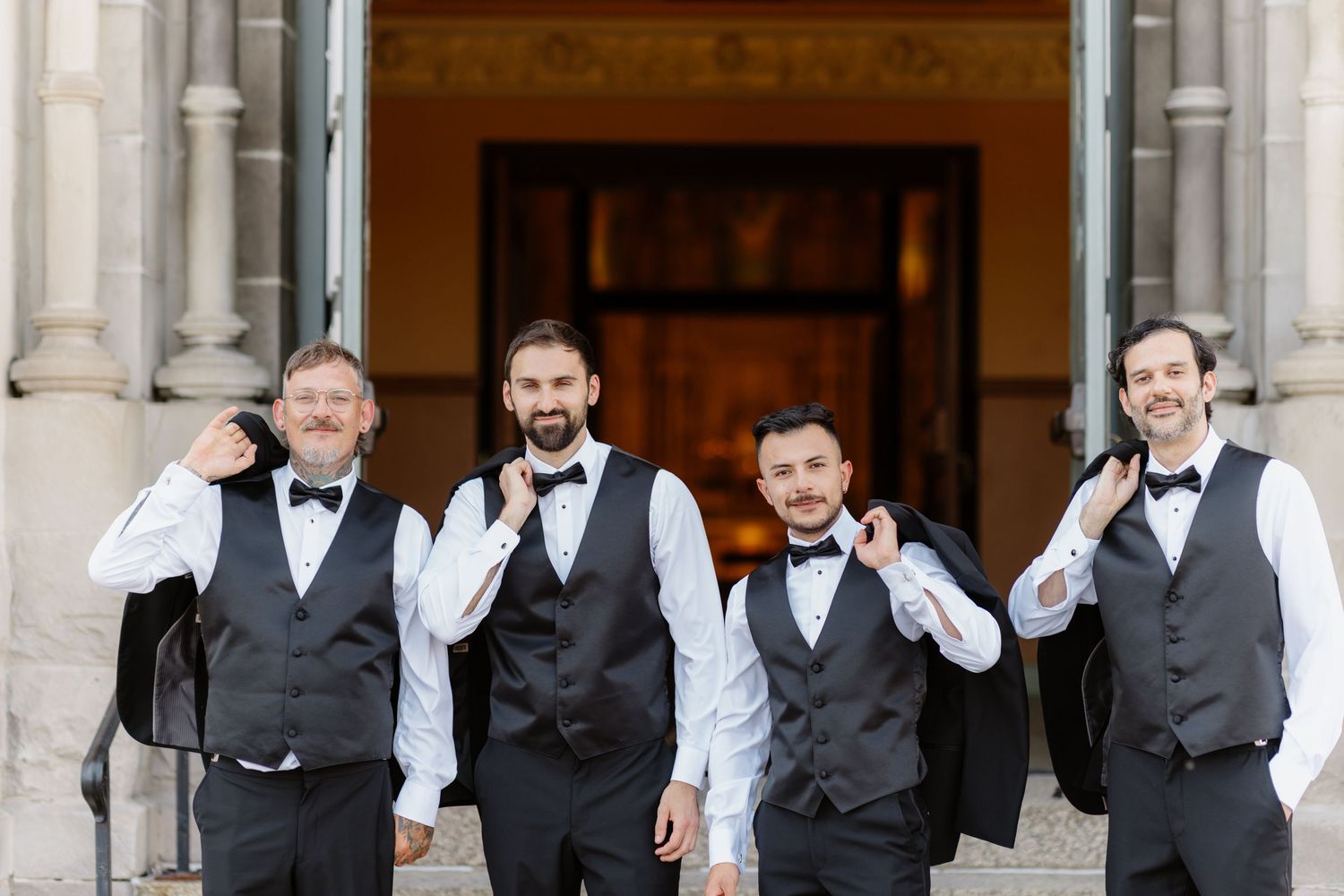 Four groomsmen in black vests and bow ties smiling and gesturing outside church entrance.