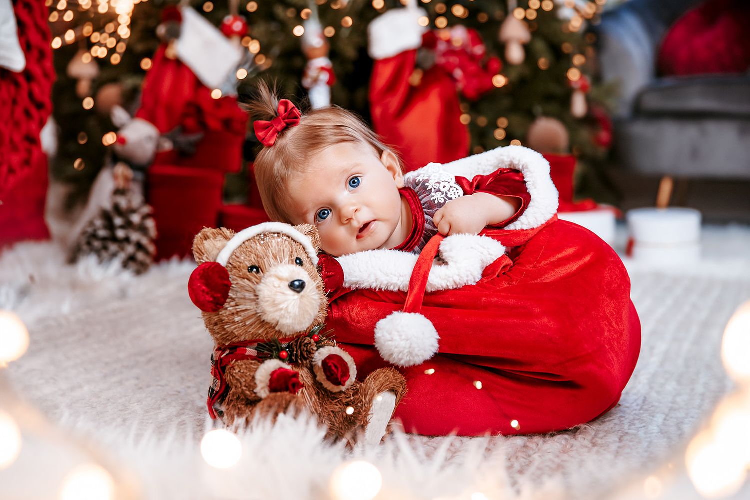 Baby girl in red Santa outfit cuddles a teddy bear beside a decorated Christmas tree with festive lights.