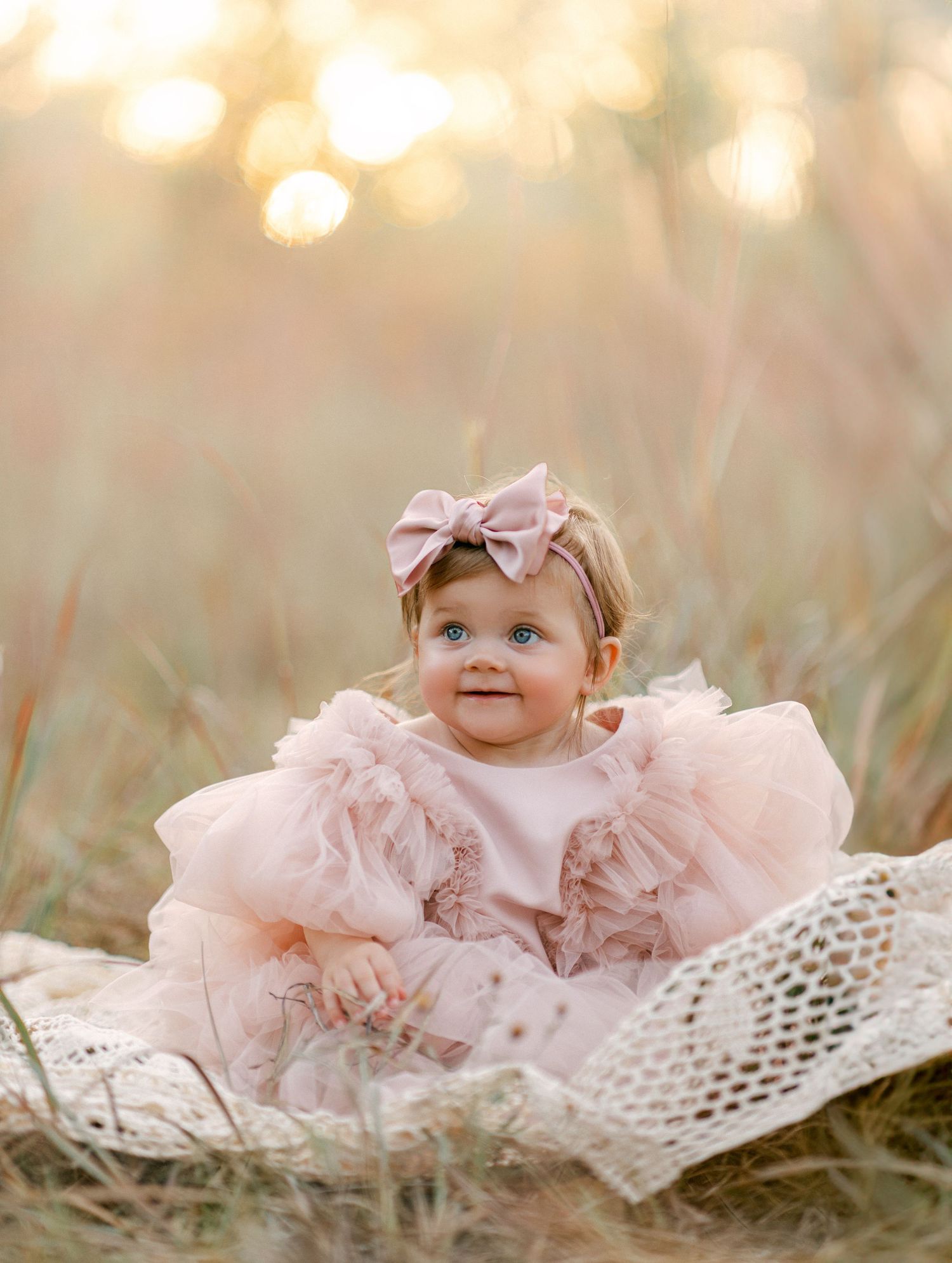 Adorable baby girl in pink tutu dress and bow headband sitting in golden field during sunset photography session.