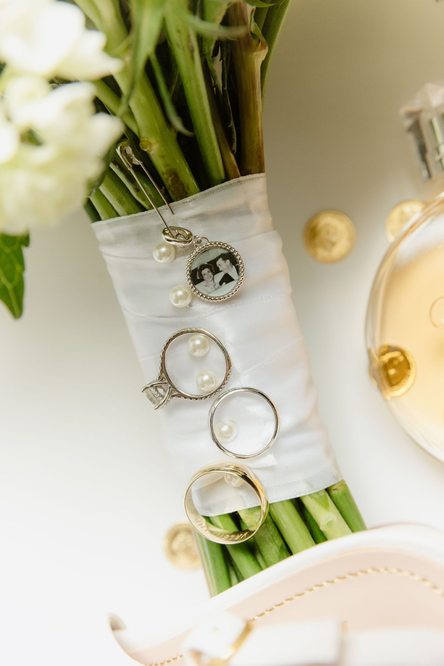 Wedding rings, pearl jewelry, and a photo charm rest on a bridal bouquet of white flowers beside perfume.