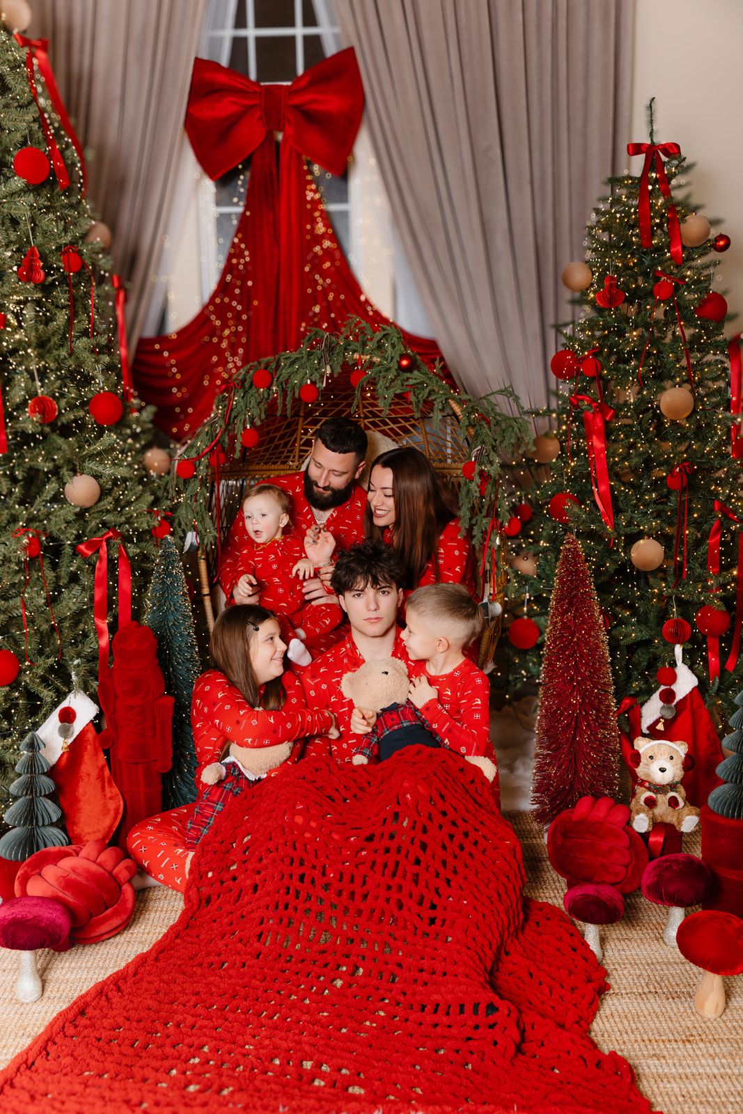Family in matching red pajamas celebrates Christmas surrounded by decorated trees, red ornaments, and festive holiday decor.