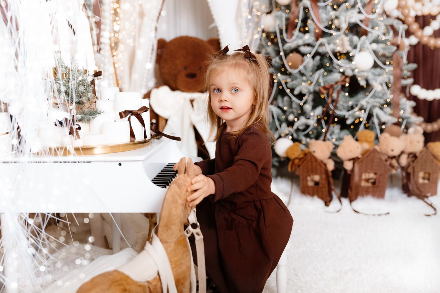 Young girl in brown dress playing white piano near decorated Christmas tree with teddy bear and holiday ornaments.