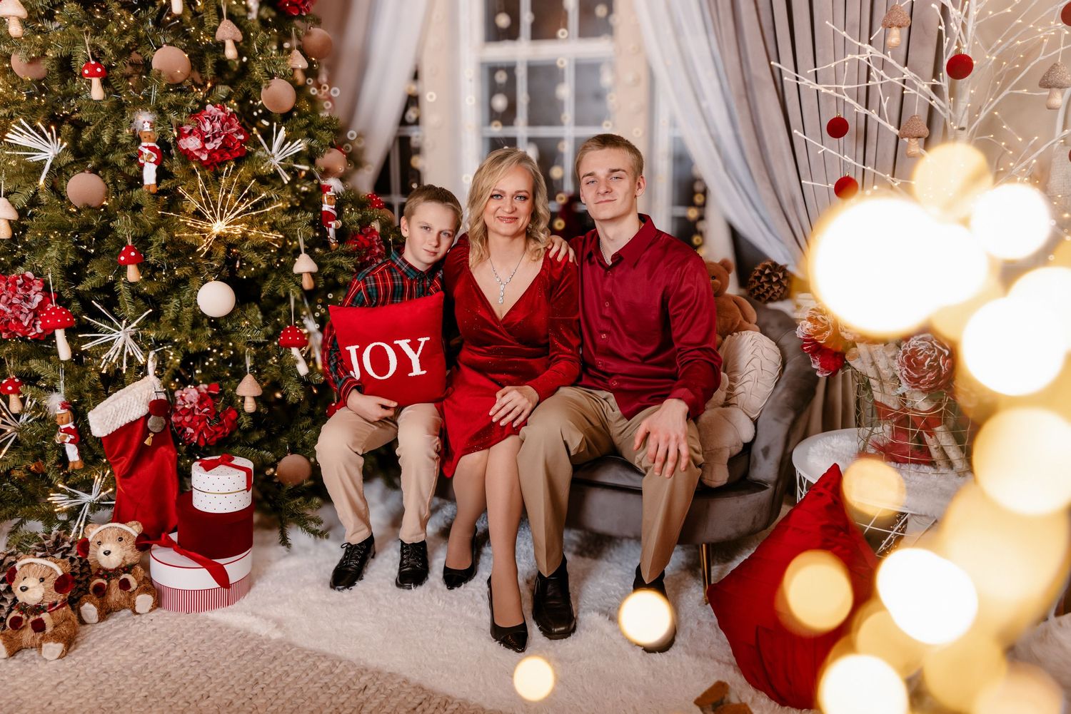 A family of three in red outfits sits together on a sofa beside a decorated Christmas tree holding a JOY pillow.