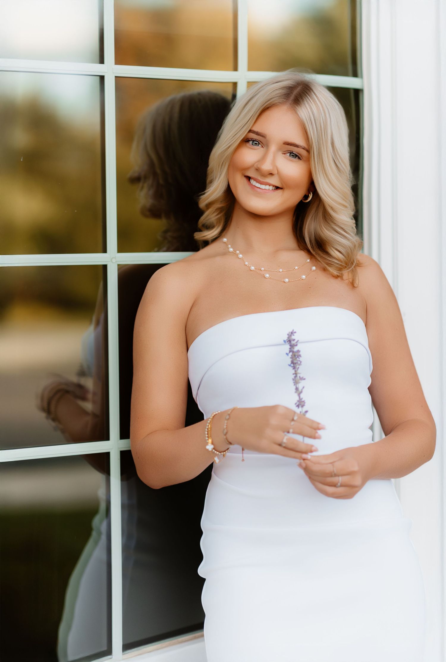 Smiling blonde woman in white strapless dress holding lavender by sunlit window.