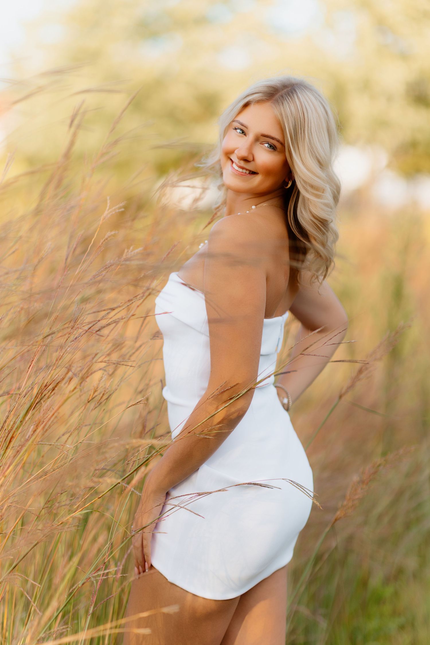 Blonde woman in white strapless dress posing in sun-lit tall grass field.