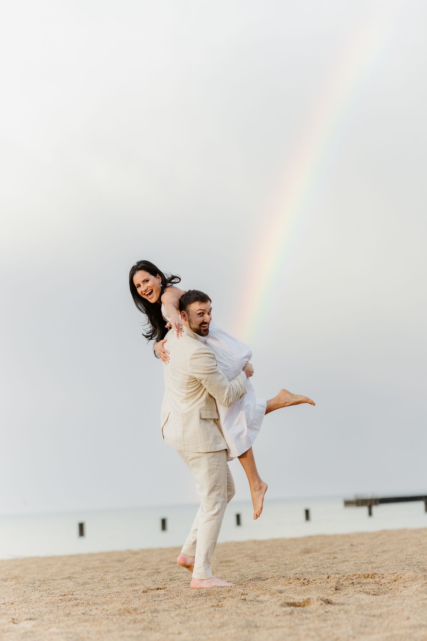 A smiling couple embracing on a sandy beach with a rainbow arching over the ocean behind them.