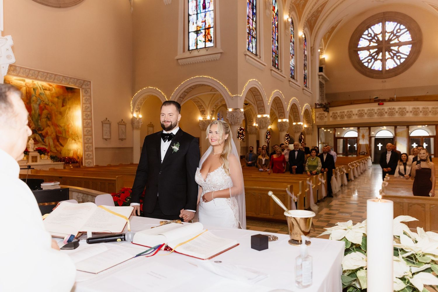 Newlyweds stand together at altar table in ornate church with arched windows and packed pews of wedding guests.